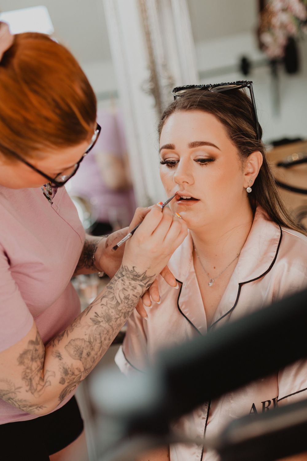 Makeup artist applying lipstick to a bride in a satin robe during bridal preparations.
