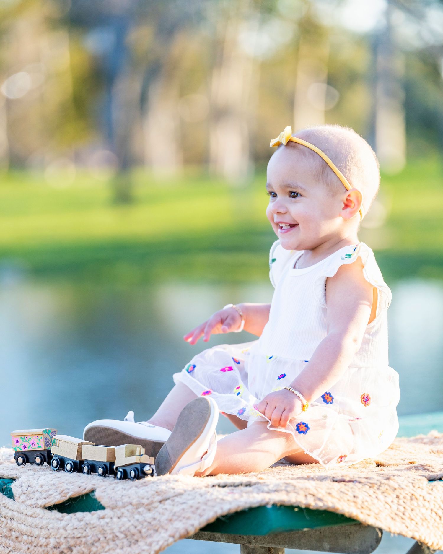 Smiling baby girl in white floral dress with yellow bow headband plays with wooden toy train outdoors near pond.