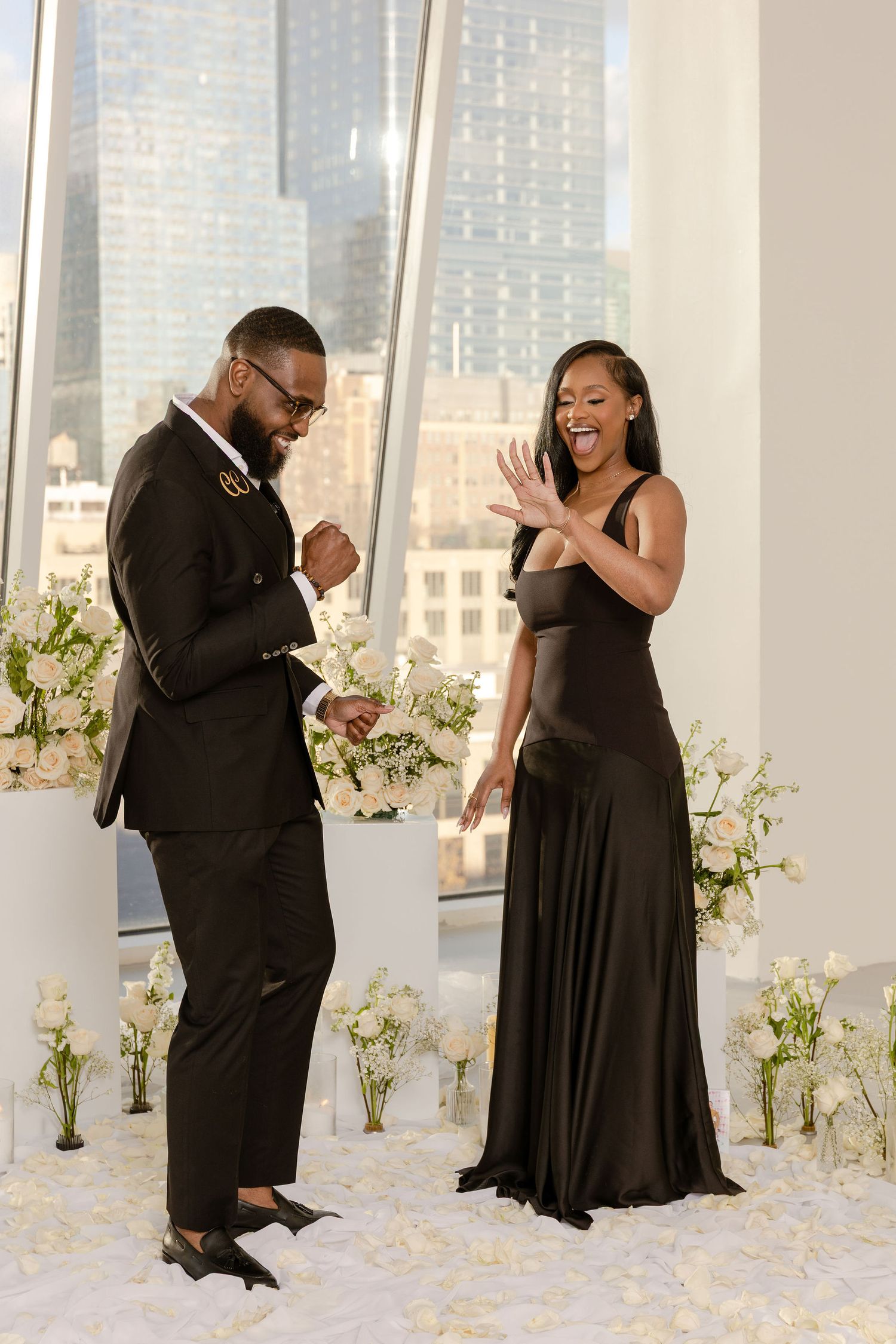 Elegant couple in black formal attire sharing a joyful moment with city skyline and white floral arrangements.