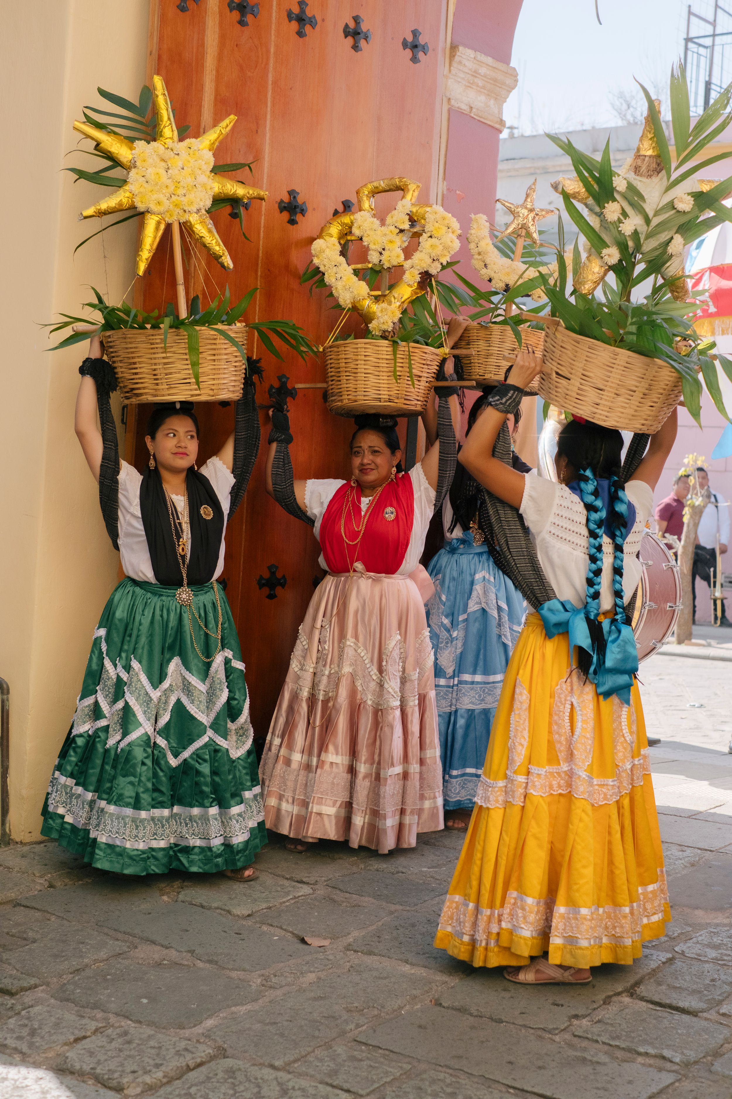 Women in traditional Mexican dresses balance flower-filled baskets on their heads during a colorful cultural festival celebration.