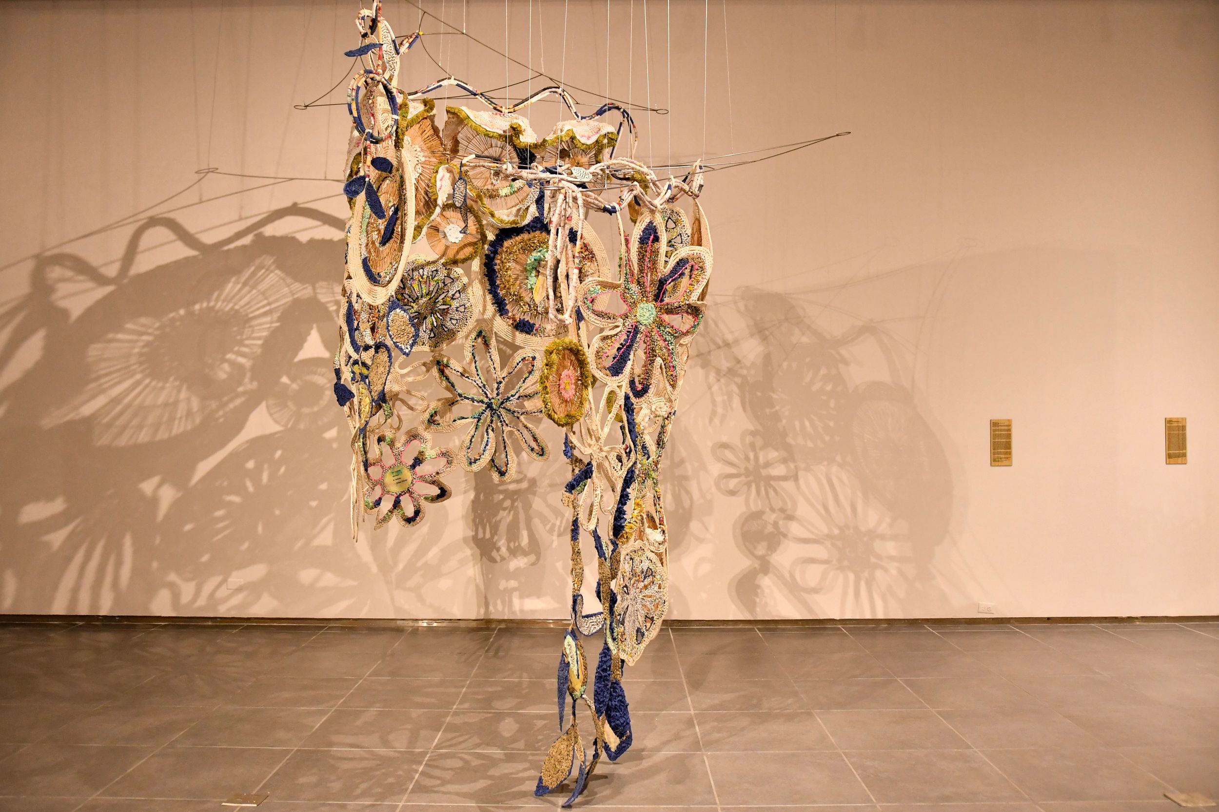 Colorful hanging sculpture with floral motifs casting intricate shadows on a gallery wall.