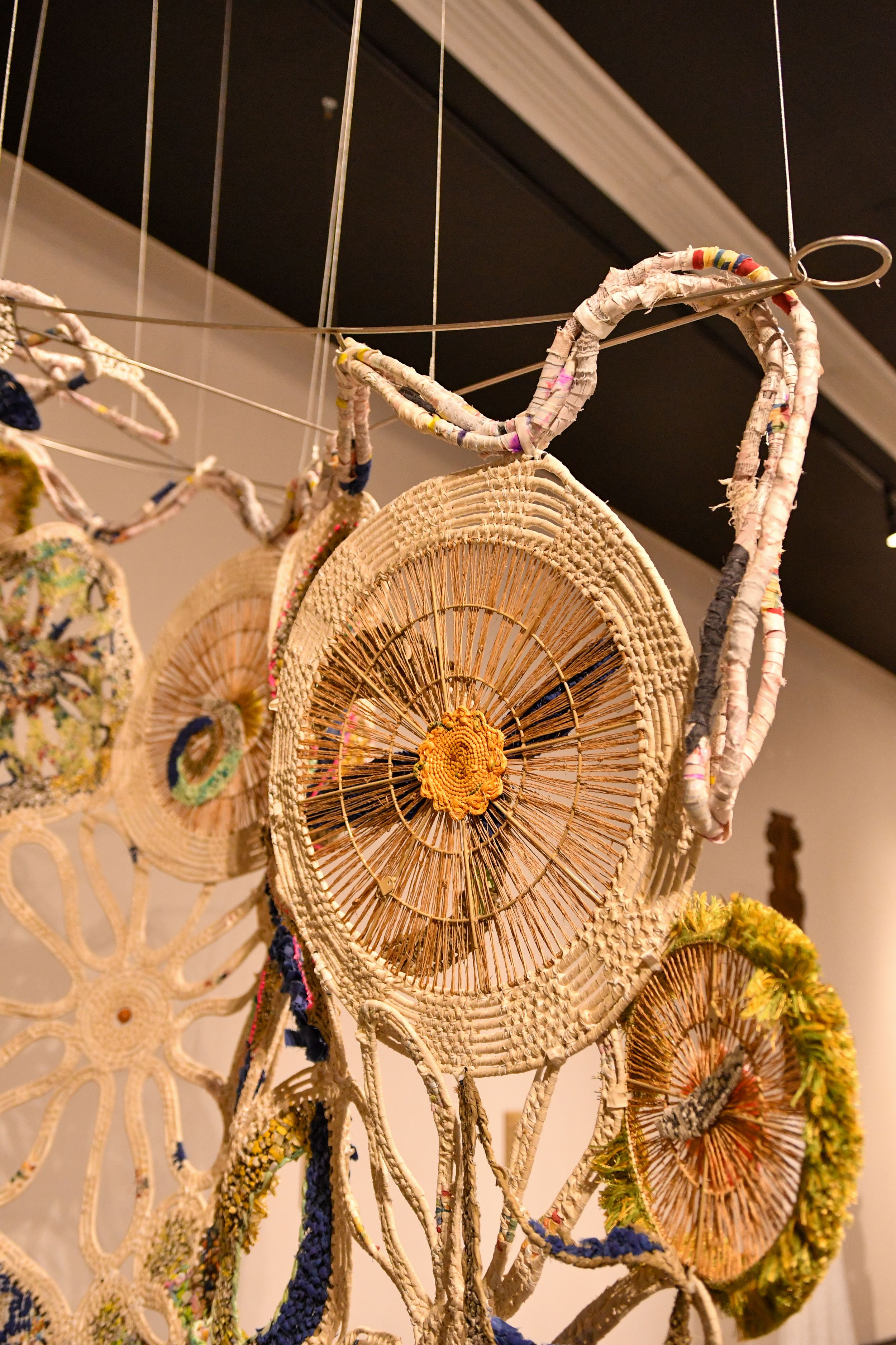 Handwoven straw and fabric dreamcatchers hanging from ceiling in an art gallery display.