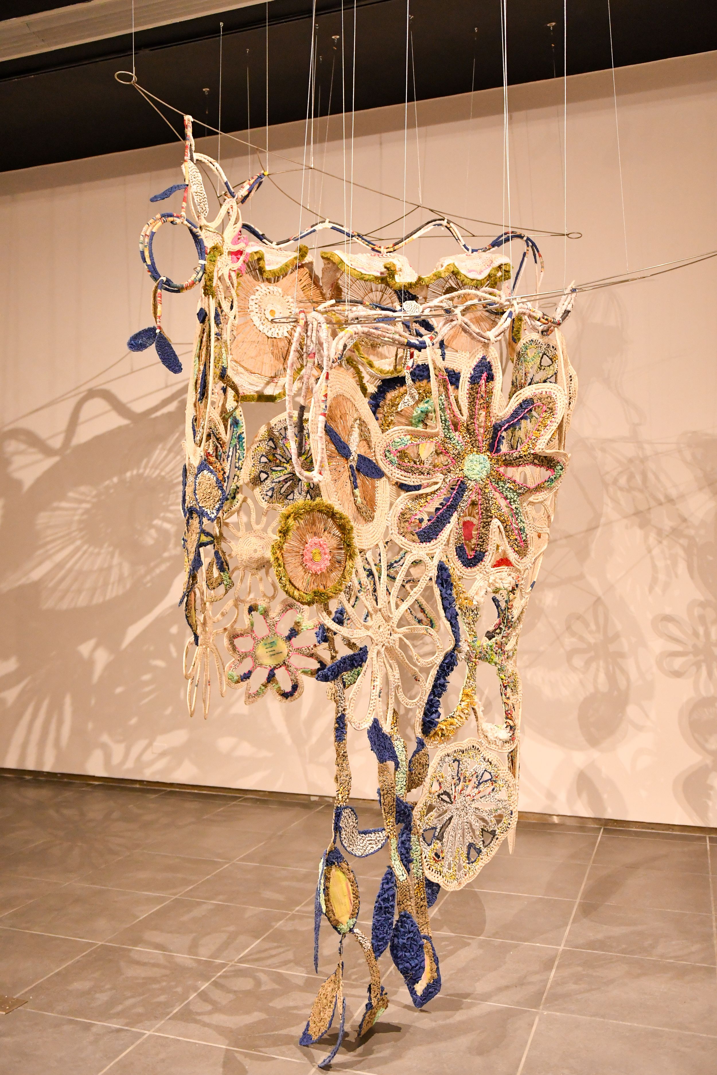 Colorful hanging textile art sculpture with floral crochet patterns suspended from wires in a gallery.