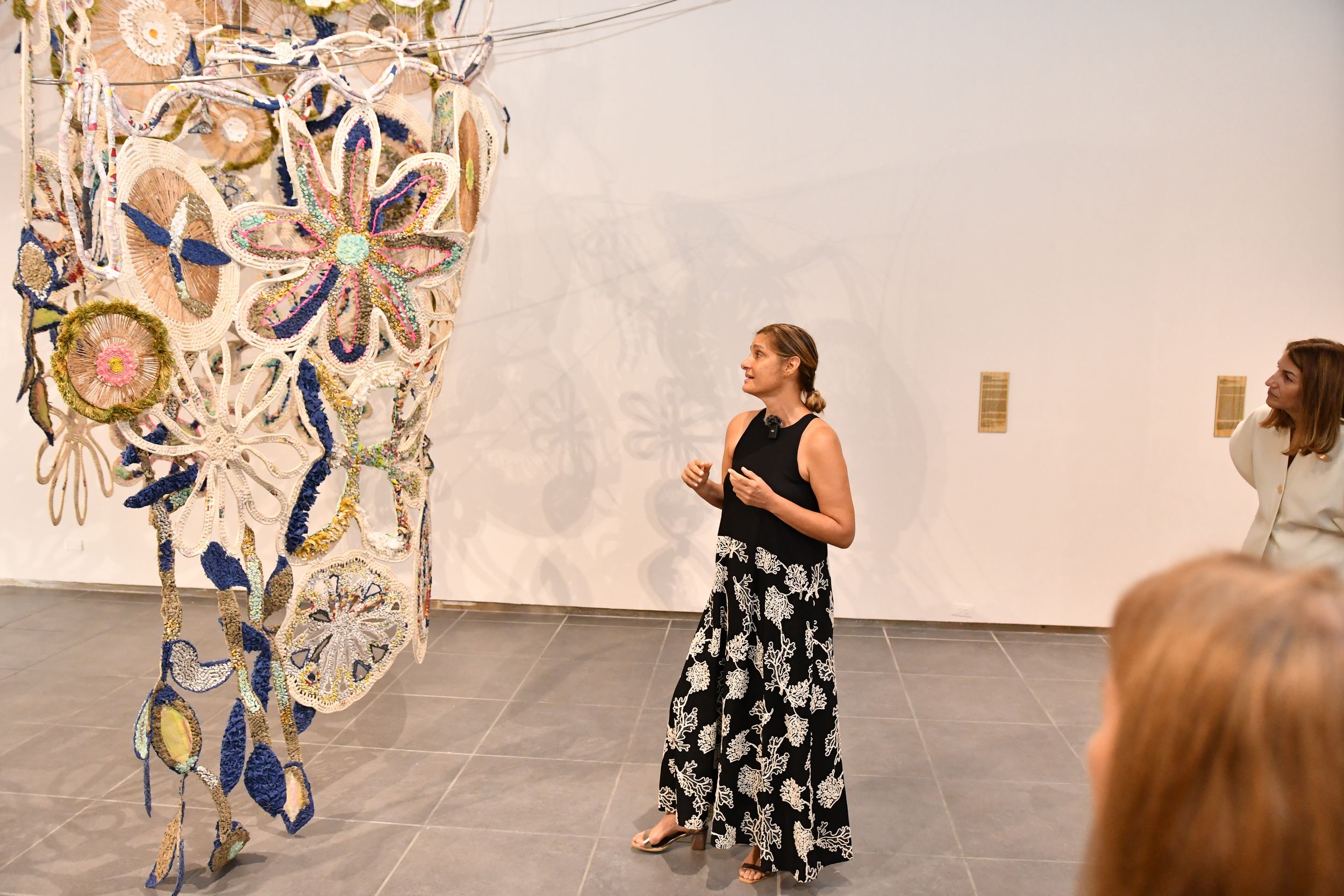 Woman in black floral dress presenting large colorful floral sculpture installation in contemporary art gallery.