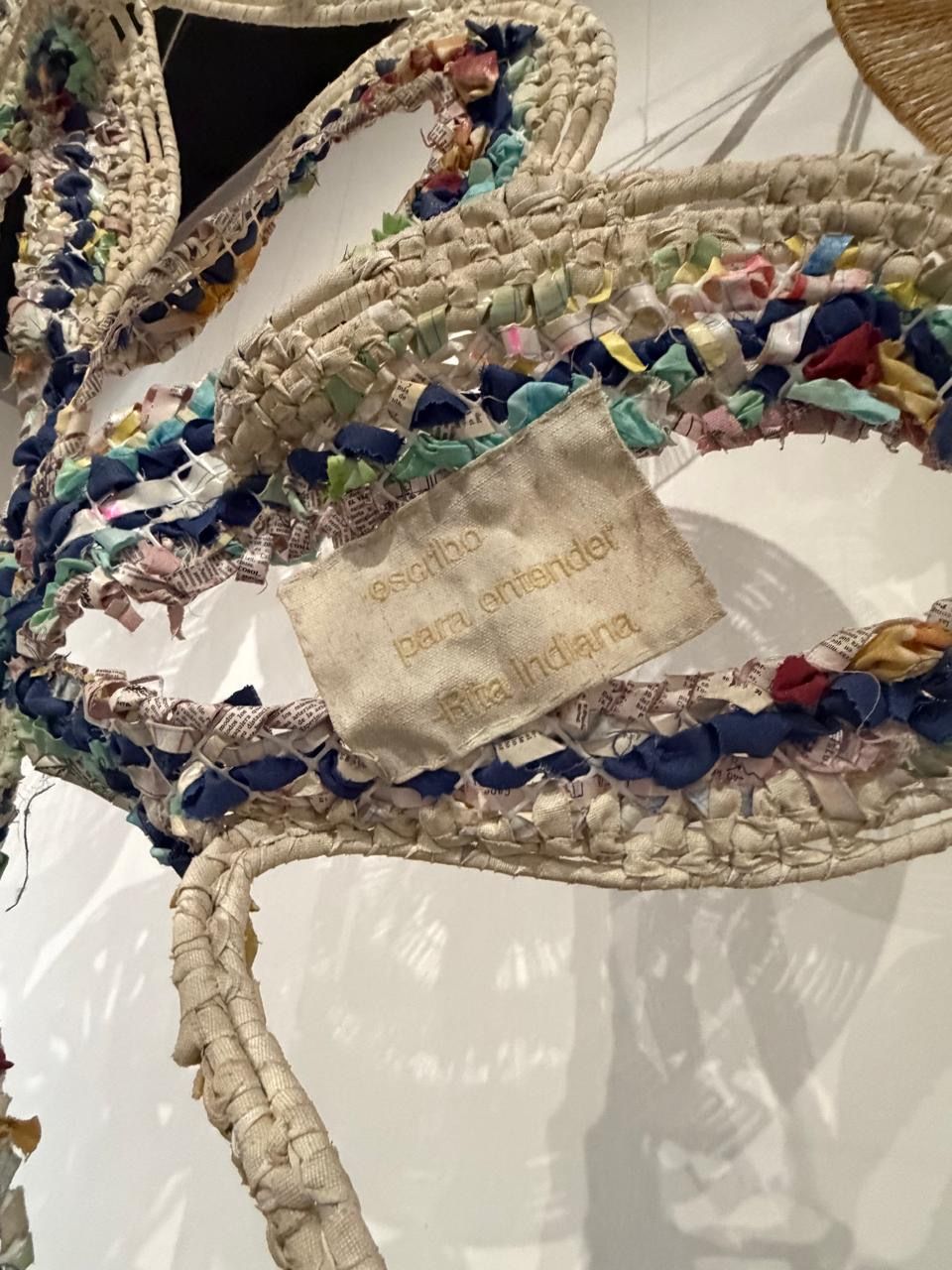 Close-up of a colorful woven textile art piece with a fabric label reading text in Spanish, displayed on a white wall.