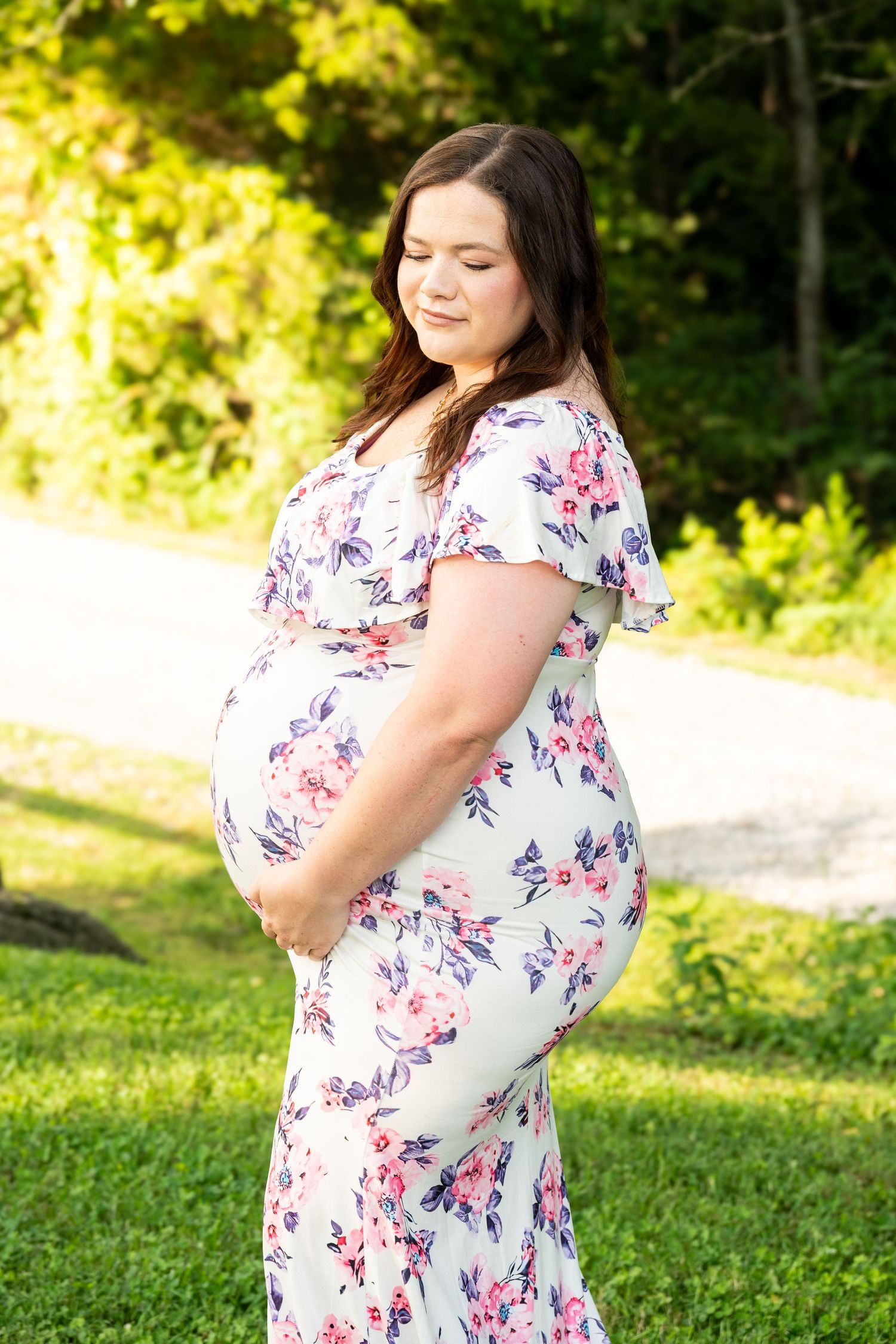 A pregnant woman in a floral dress cradles her baby bump while standing outdoors in natural light.