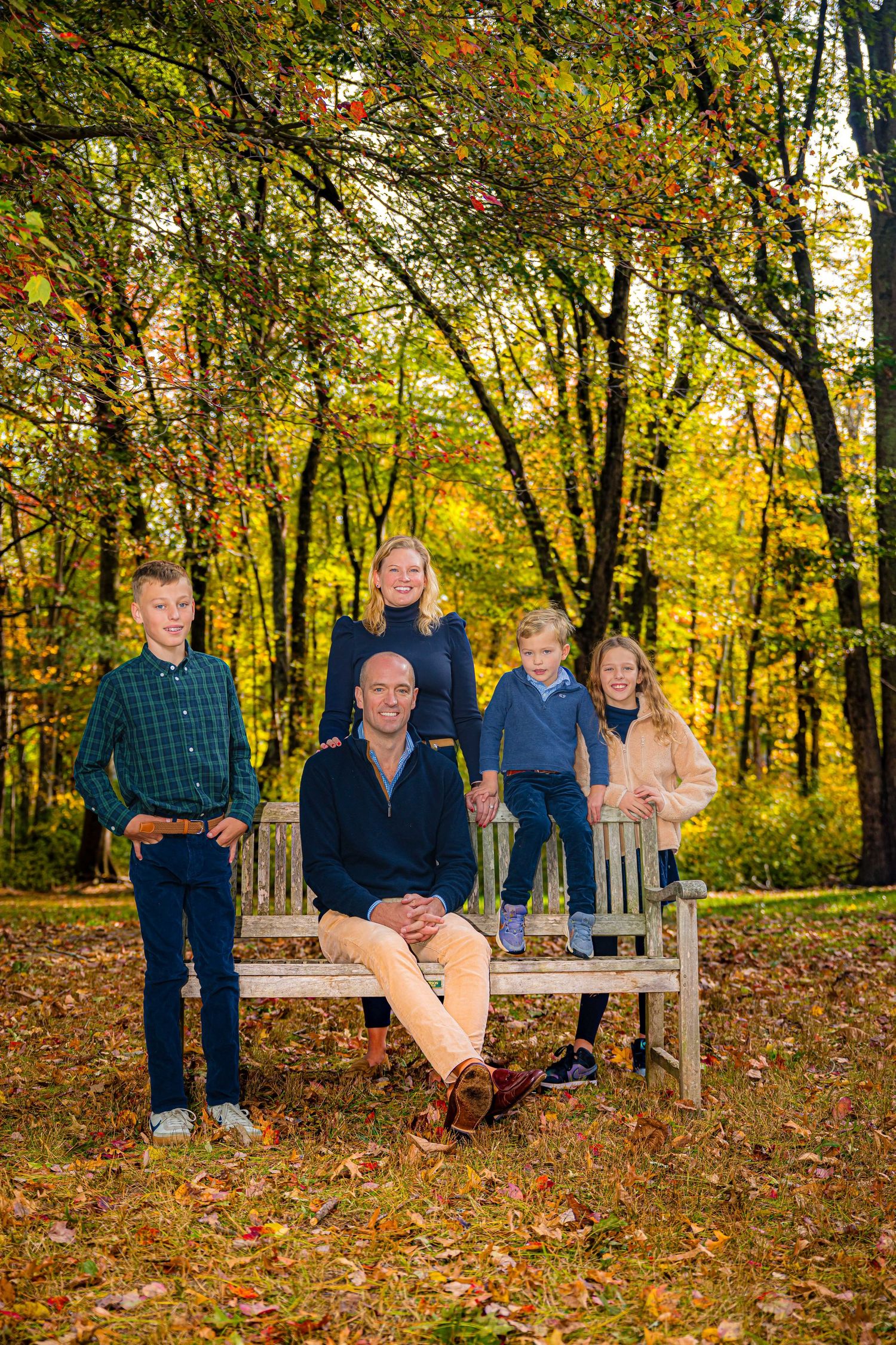 Family of five posing on a wooden bench surrounded by colorful autumn foliage in a scenic park setting.