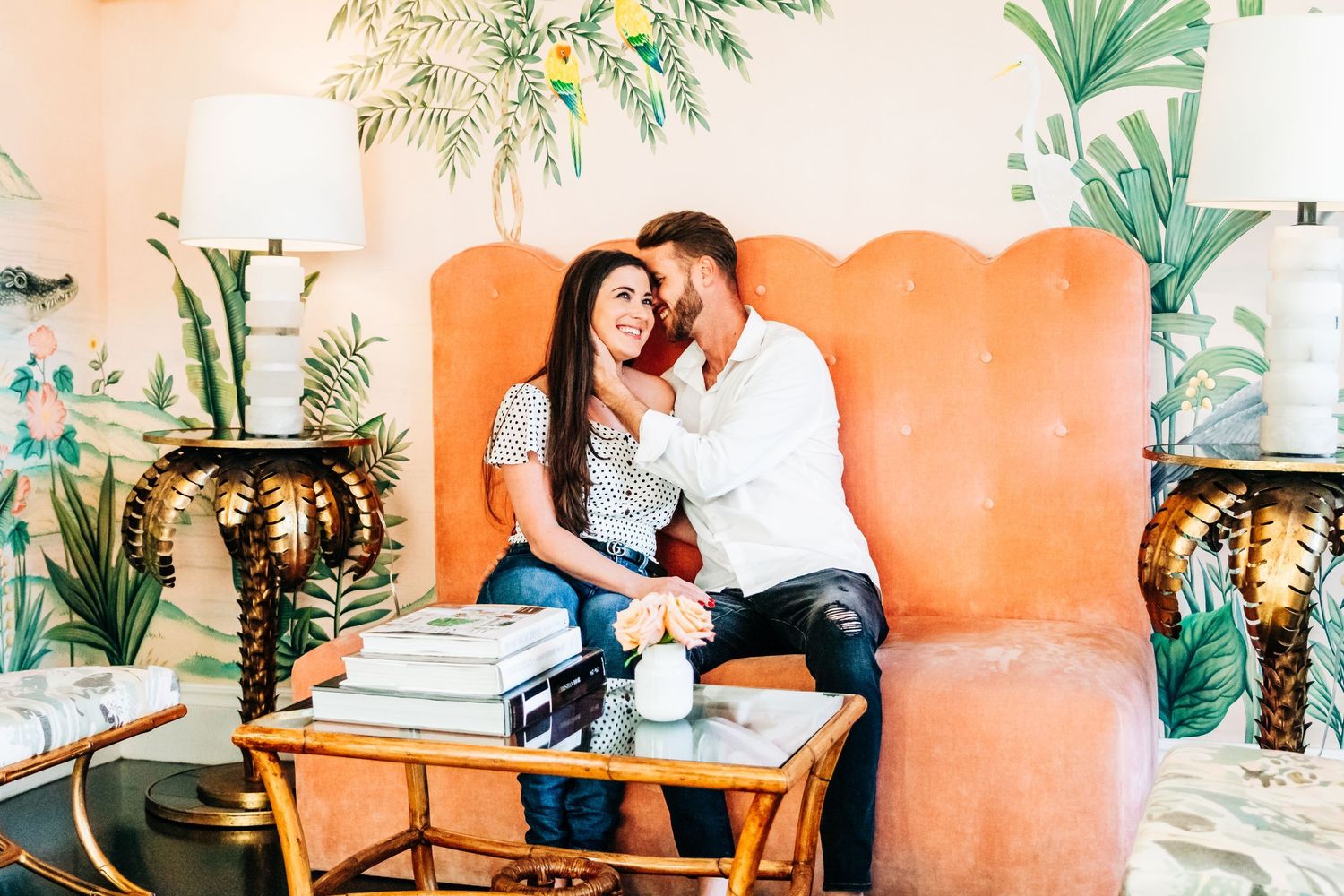 Couple sitting together on an orange velvet chair in a tropical themed room with palm leaf wallpaper at historic Colony Palm Beach hotel.