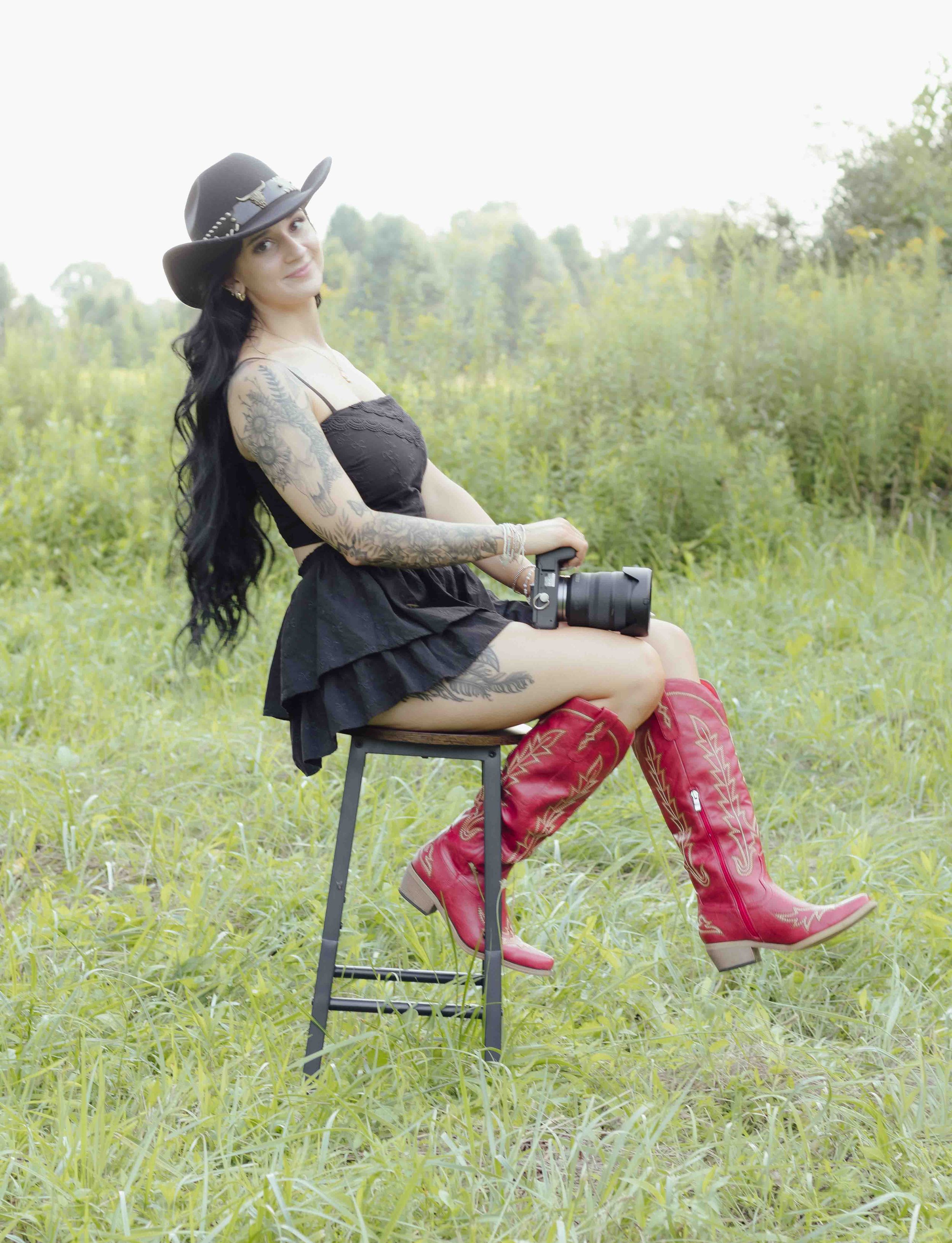 Tattooed woman in black dress, cowboy hat, and red boots sits on stool holding camera in green field.