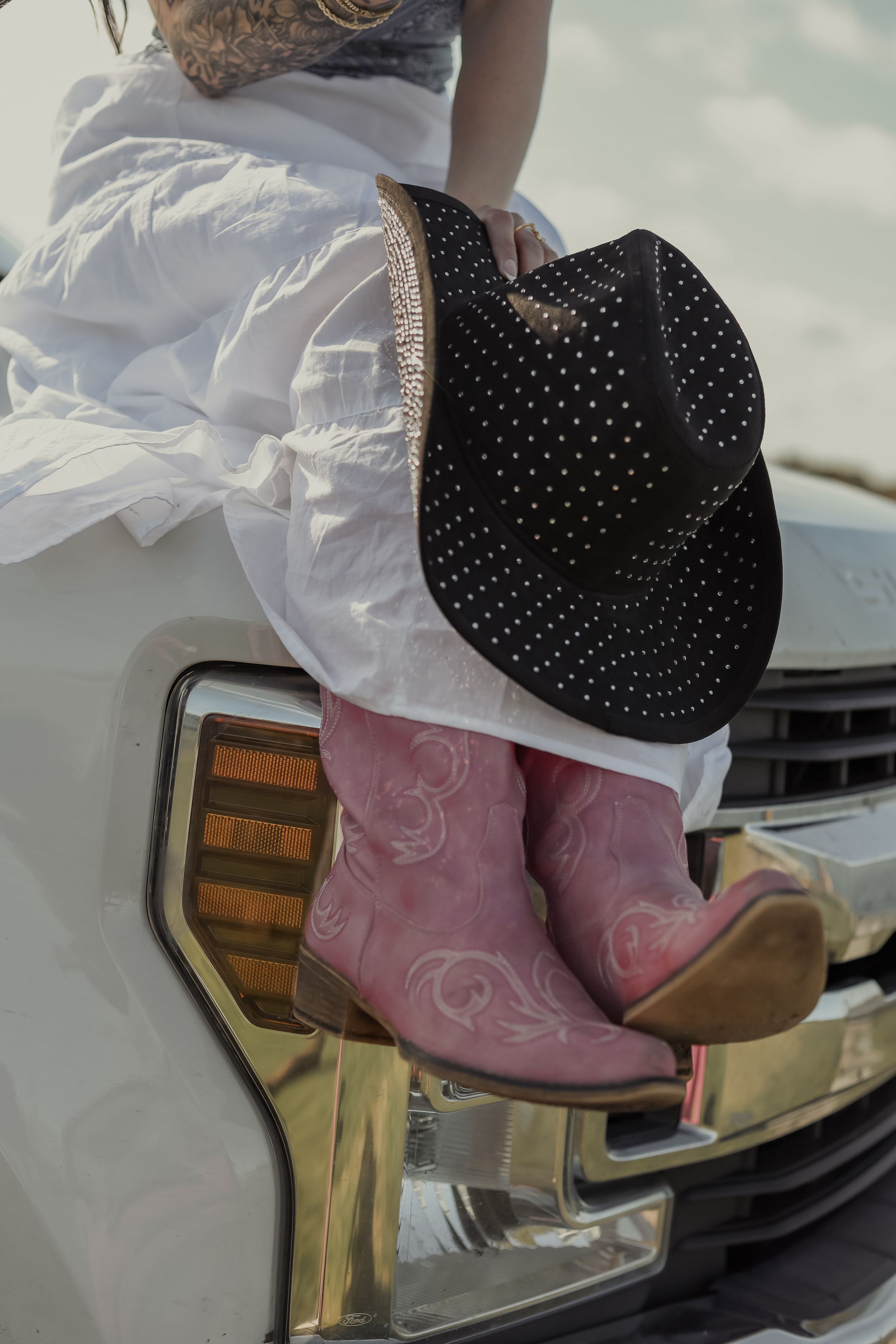 Pink cowboy boots and black studded hat on a white truck hood with a white dress.