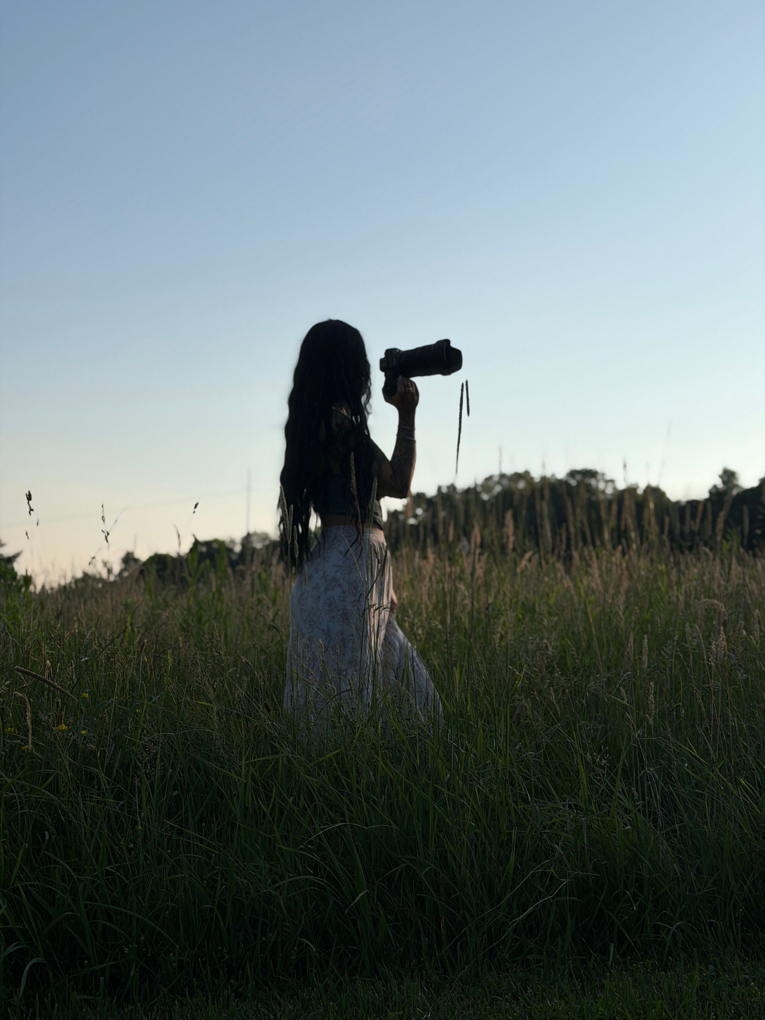 Silhouette of a woman with long hair holding a camera in a lush green field at dusk.