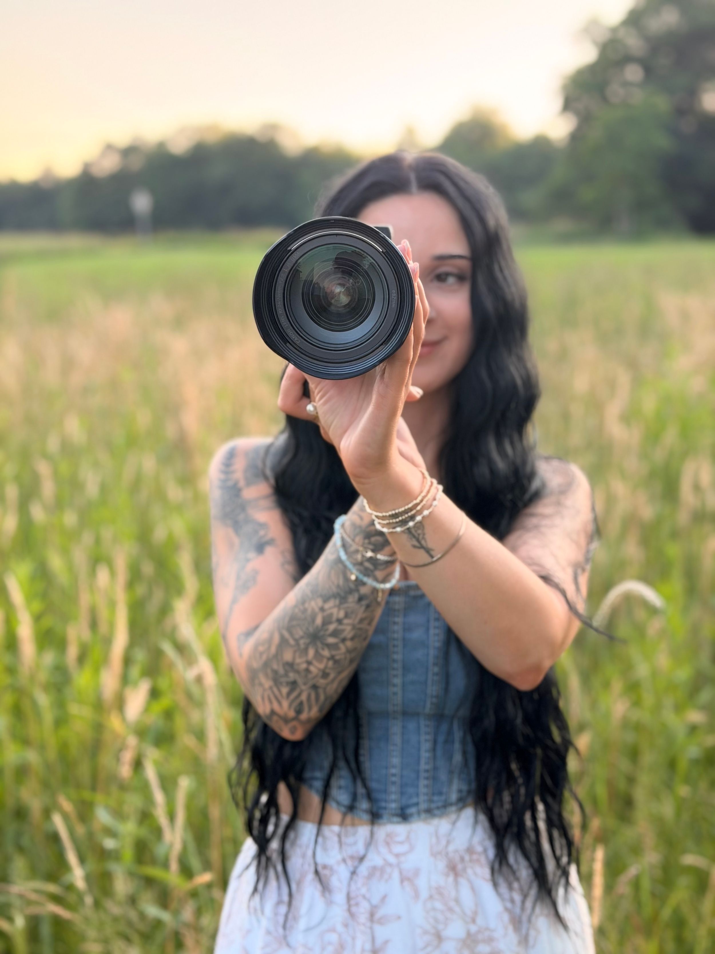 Tattooed woman with long dark hair holds a DSLR camera lens up in a sunny green field at golden hour.