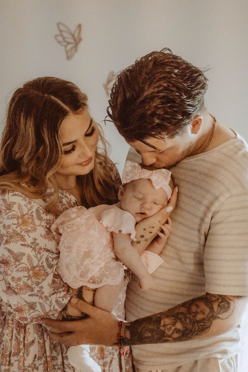 A couple lovingly holds their newborn baby dressed in pink, with the father gently kissing the infant's head.