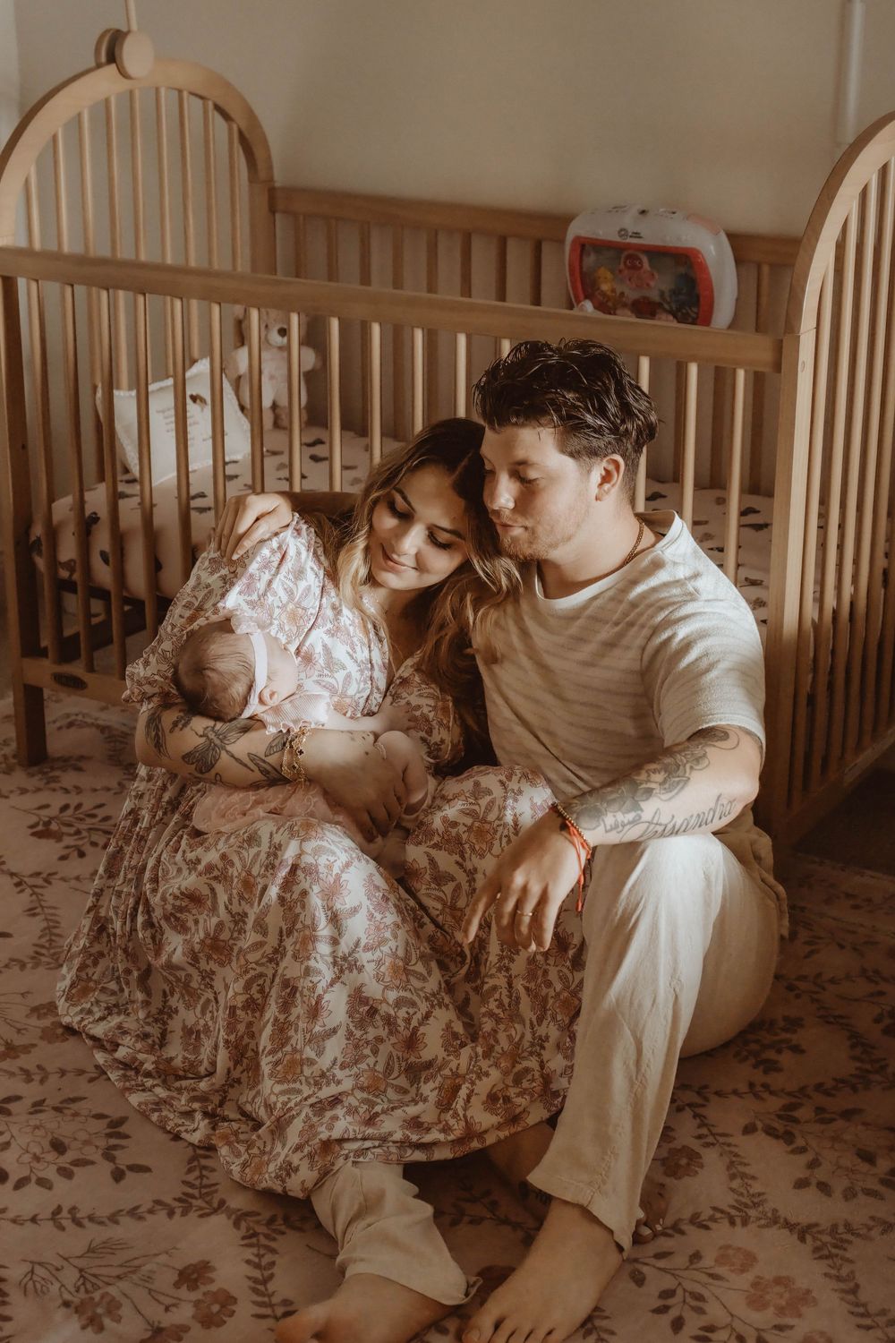 A couple sits on a floral rug beside a wooden crib, tenderly cradling their newborn baby in a warm nursery.