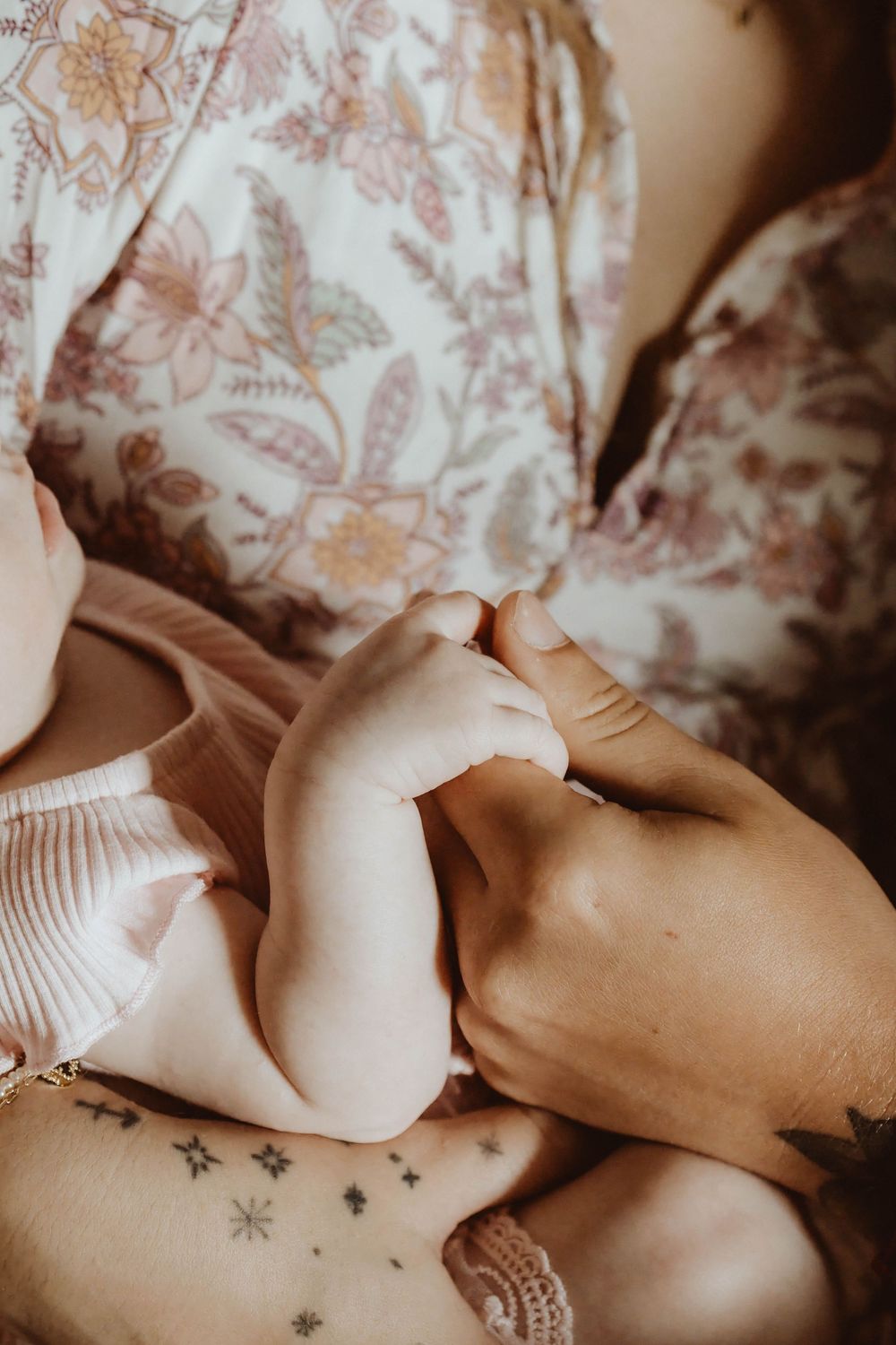Close-up of a mother in floral dress cradling her newborn baby's tiny feet, with star tattoos visible on her wrist.