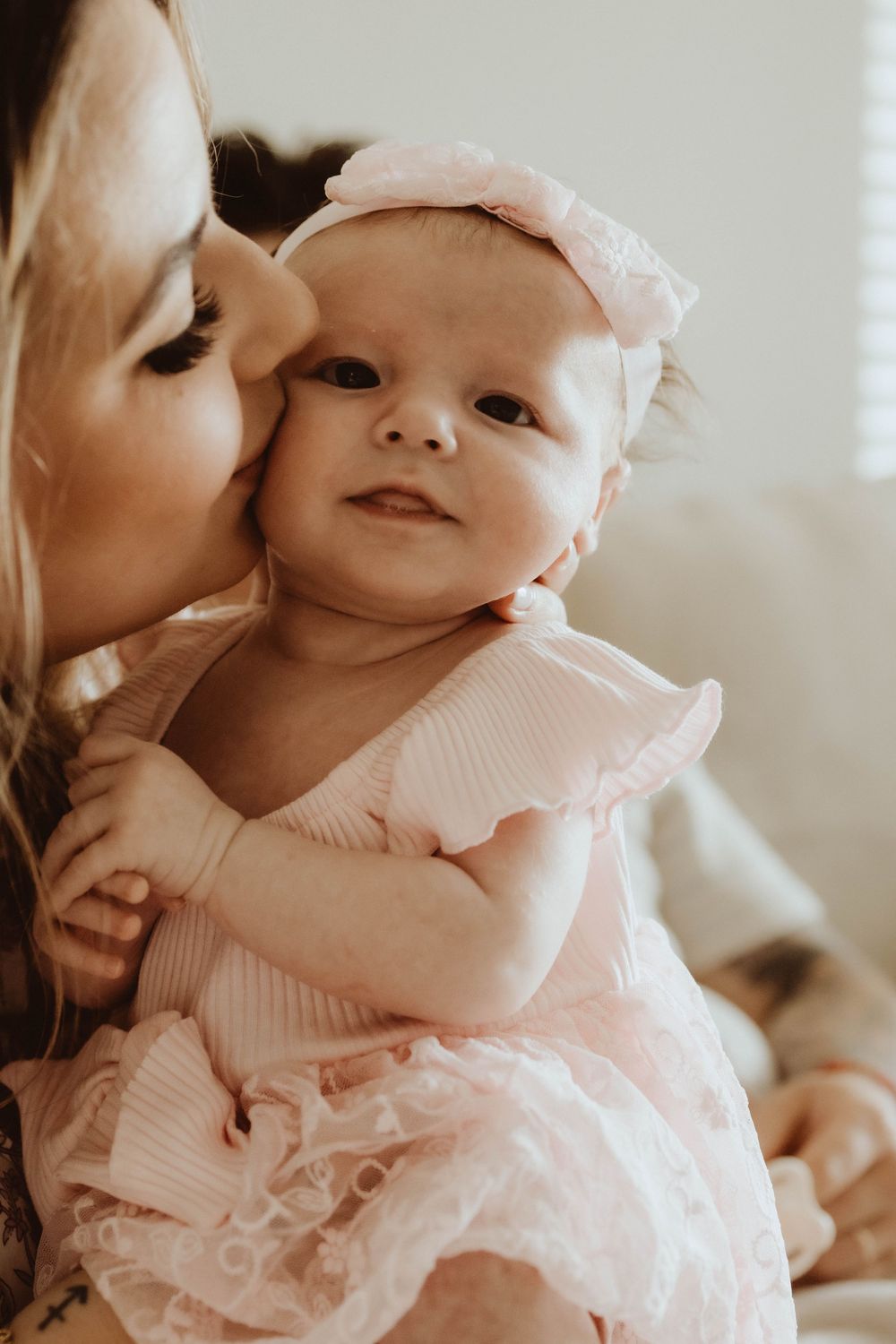 A mother tenderly kisses her smiling baby girl dressed in a pink ruffle outfit with a matching bow headband.