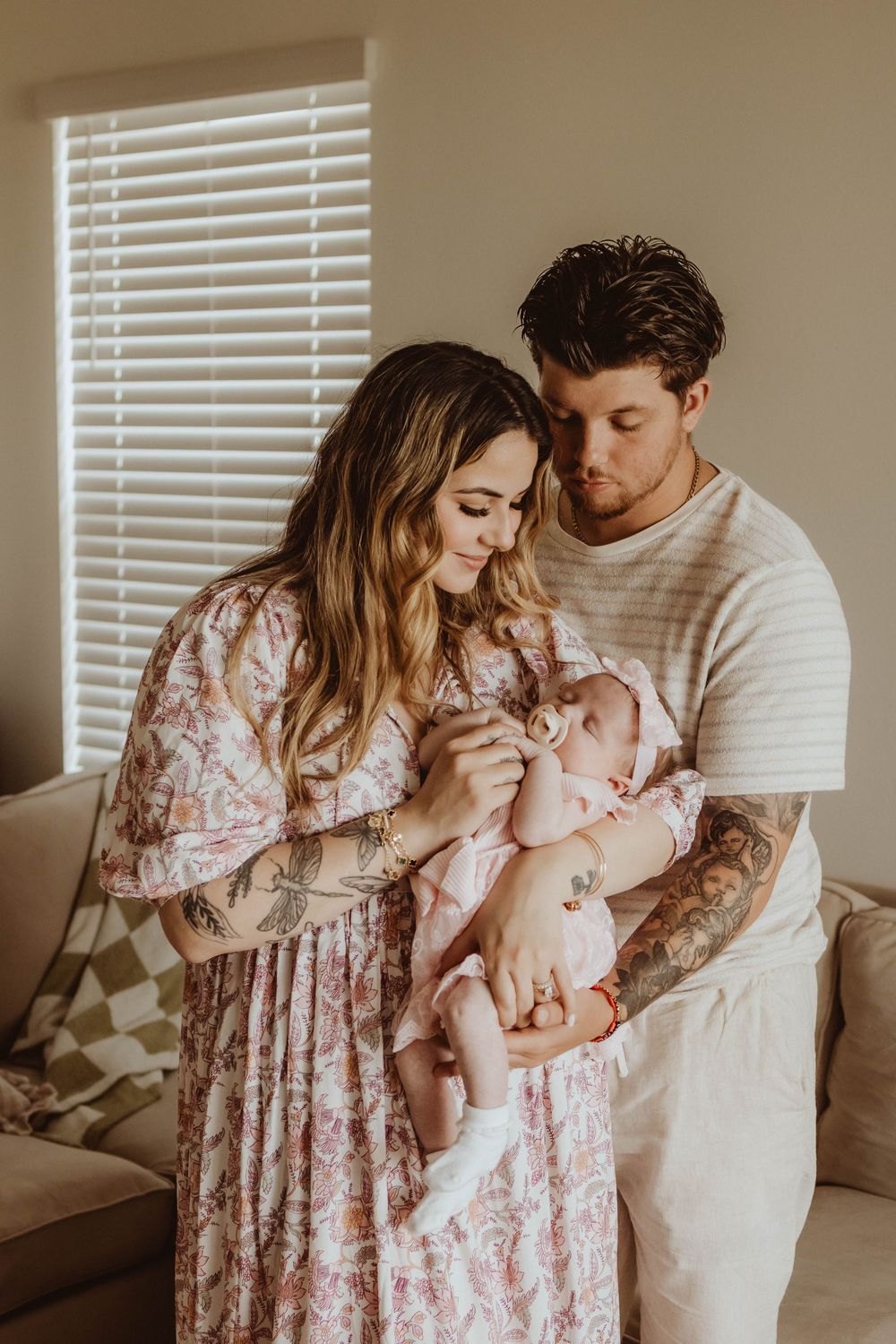 A young couple with tattoos lovingly gazes at their newborn baby indoors near a window with white blinds.