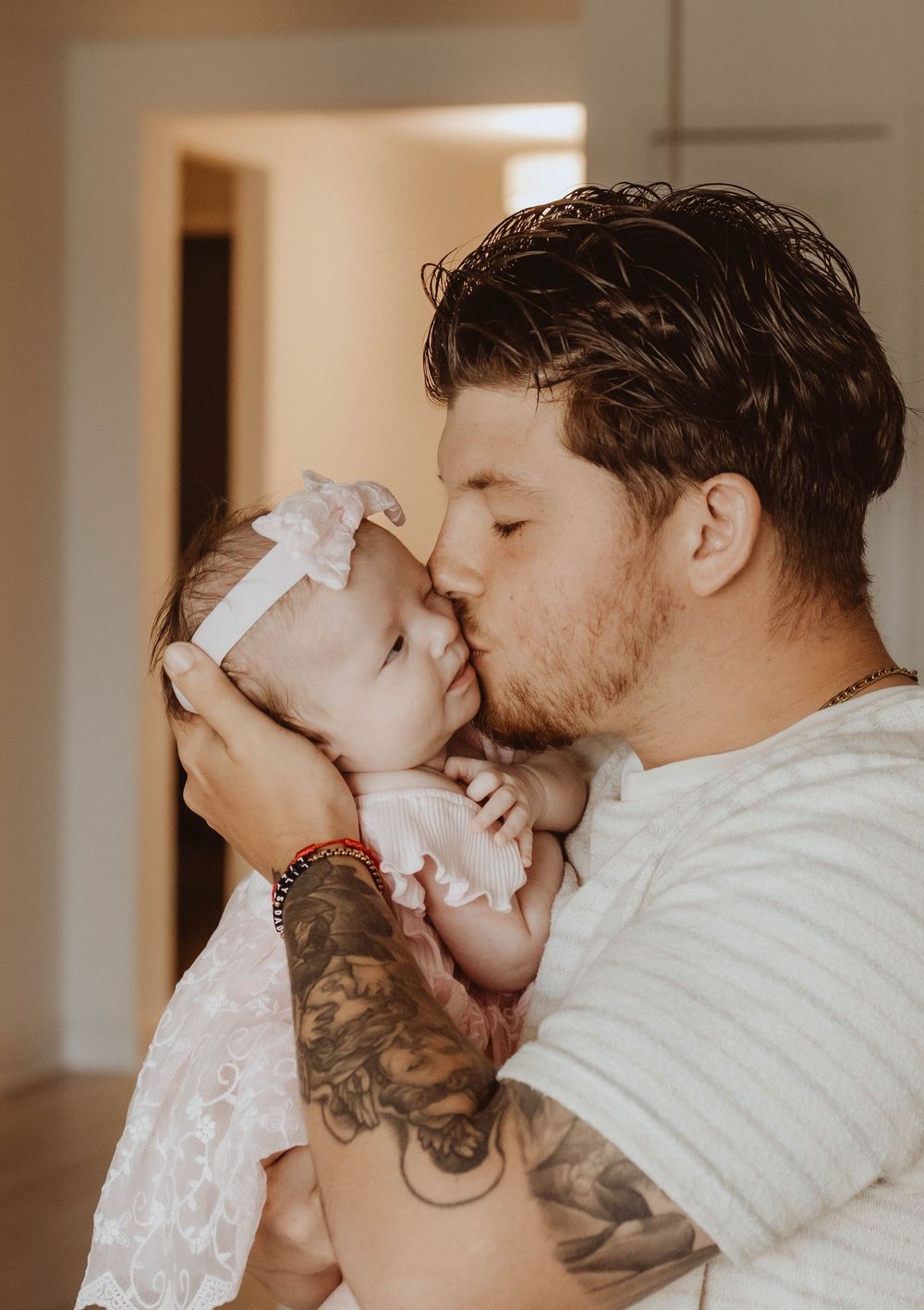 Tattooed father gently kissing his newborn baby girl wearing a white bow headband indoors.