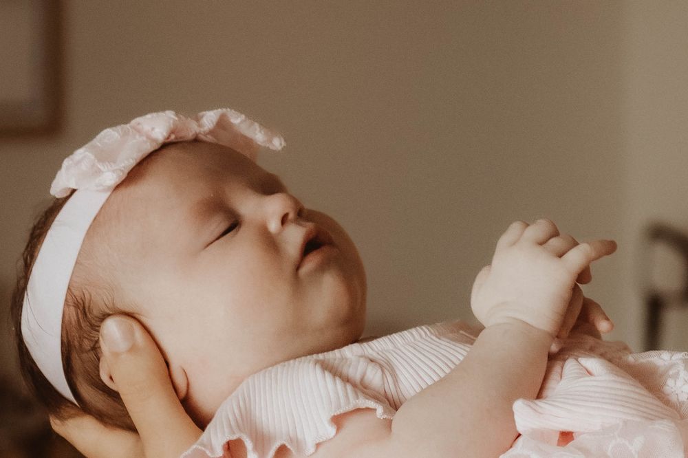 Peaceful newborn baby girl sleeping in pink outfit with white bow headband, eyes closed, tiny hands raised.