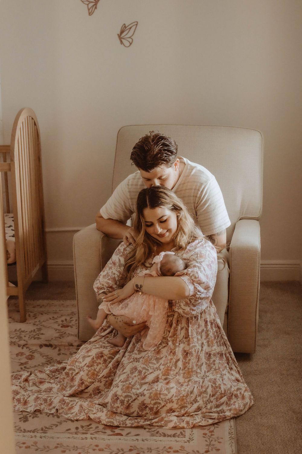 A couple sits in a nursery chair, lovingly gazing at their newborn baby, surrounded by soft neutral decor.