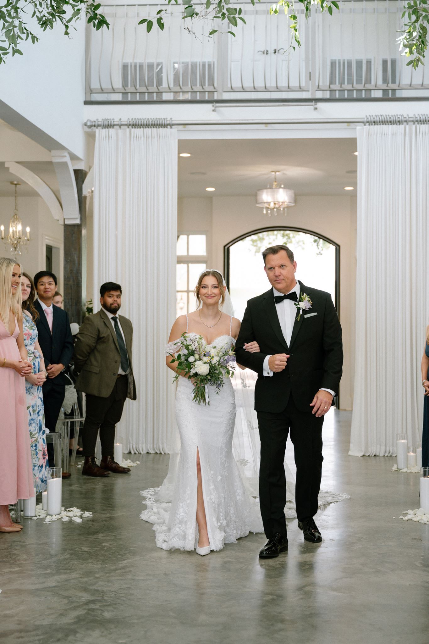 Bride in lace gown walks down aisle with escort in tuxedo at elegant white wedding venue with floral bouquet.