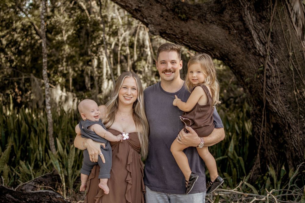 Family of four posing outdoors under a large tree, parents holding two young children, smiling in a lush green setting.