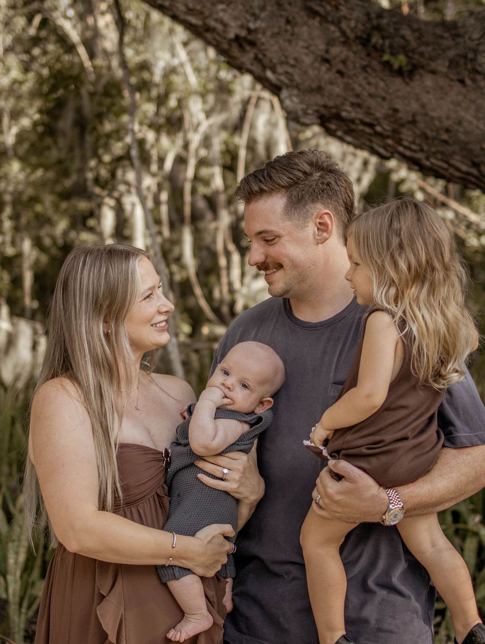 Happy family of four with parents holding a newborn baby and toddler girl outdoors under a mossy tree.