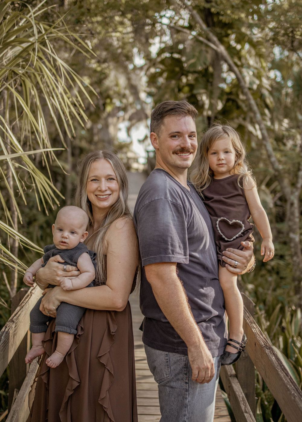 A smiling family of four poses on a wooden bridge surrounded by lush greenery, parents each holding a young child.