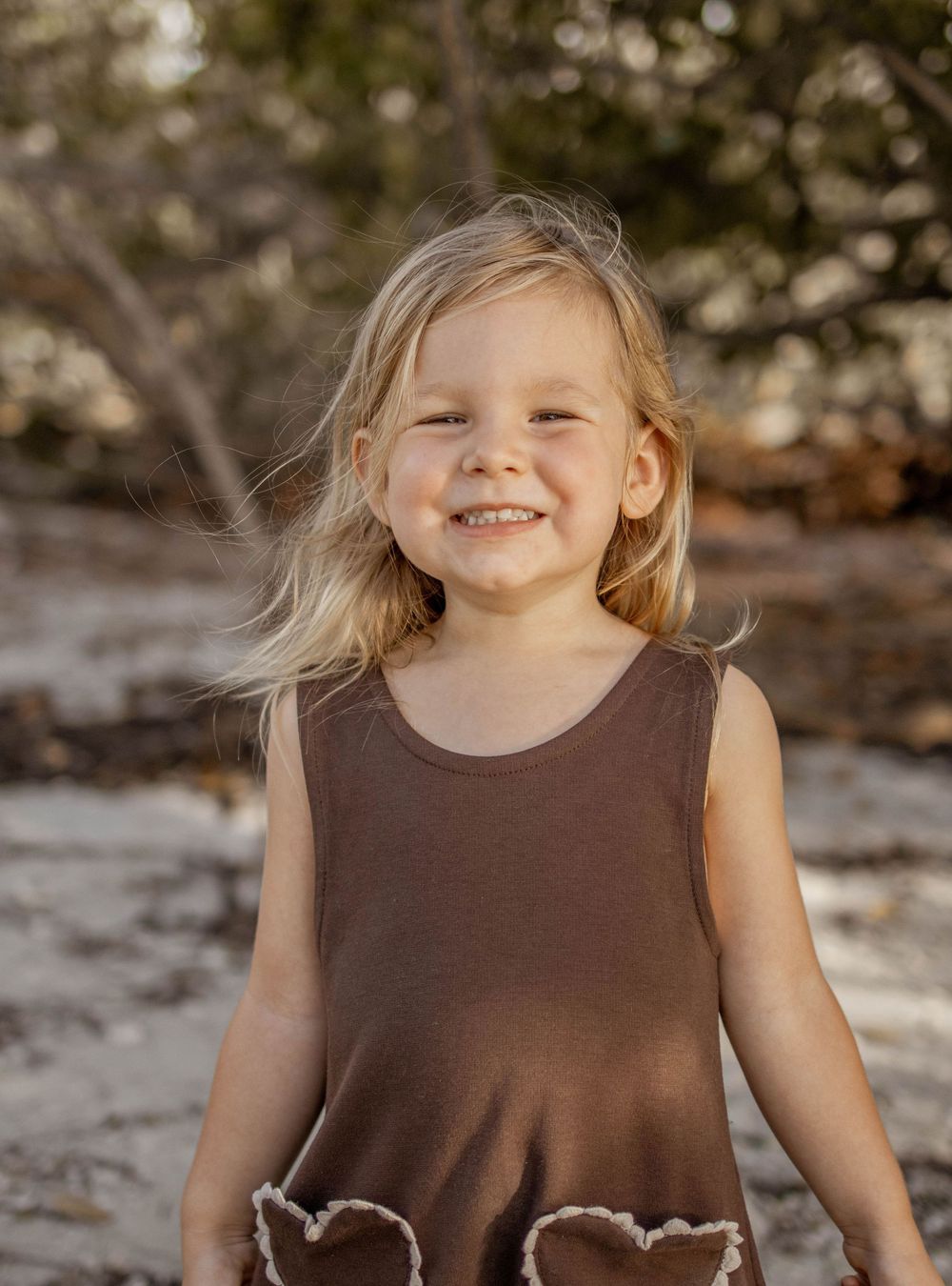 A smiling young blonde girl in a brown sleeveless dress poses outdoors with trees in the background.