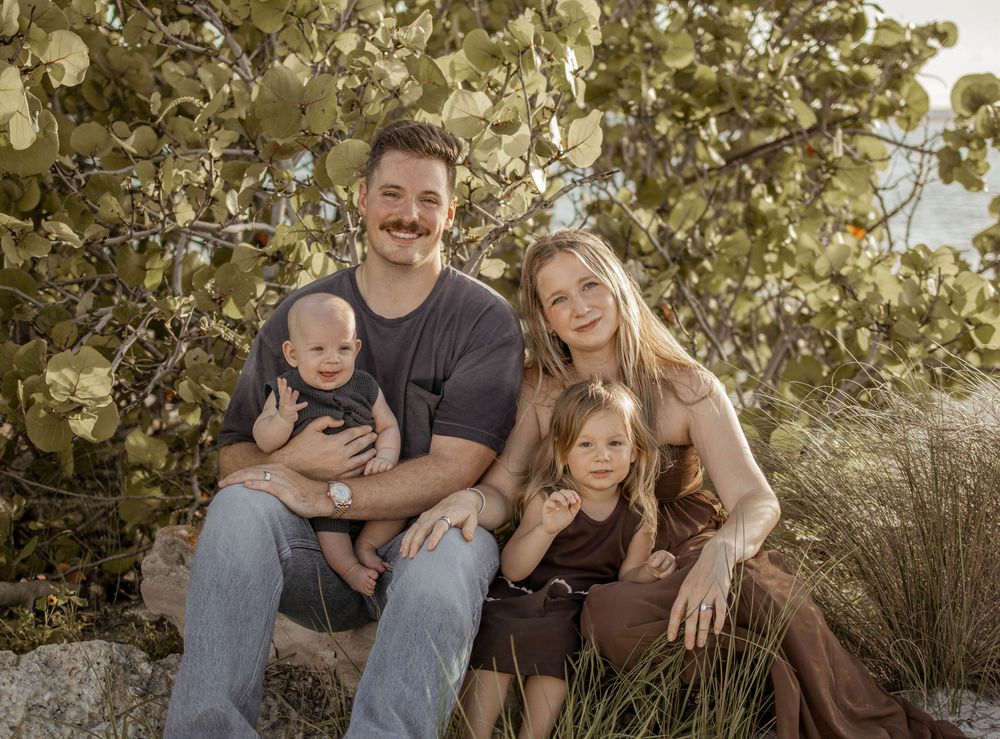 Family of four posing outdoors near lush green shrubs, parents holding young children in a warm, natural setting.