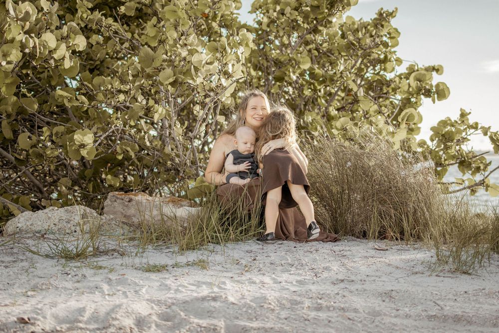 Two women sitting together on sandy beach dunes surrounded by lush green trees in a serene outdoor setting.