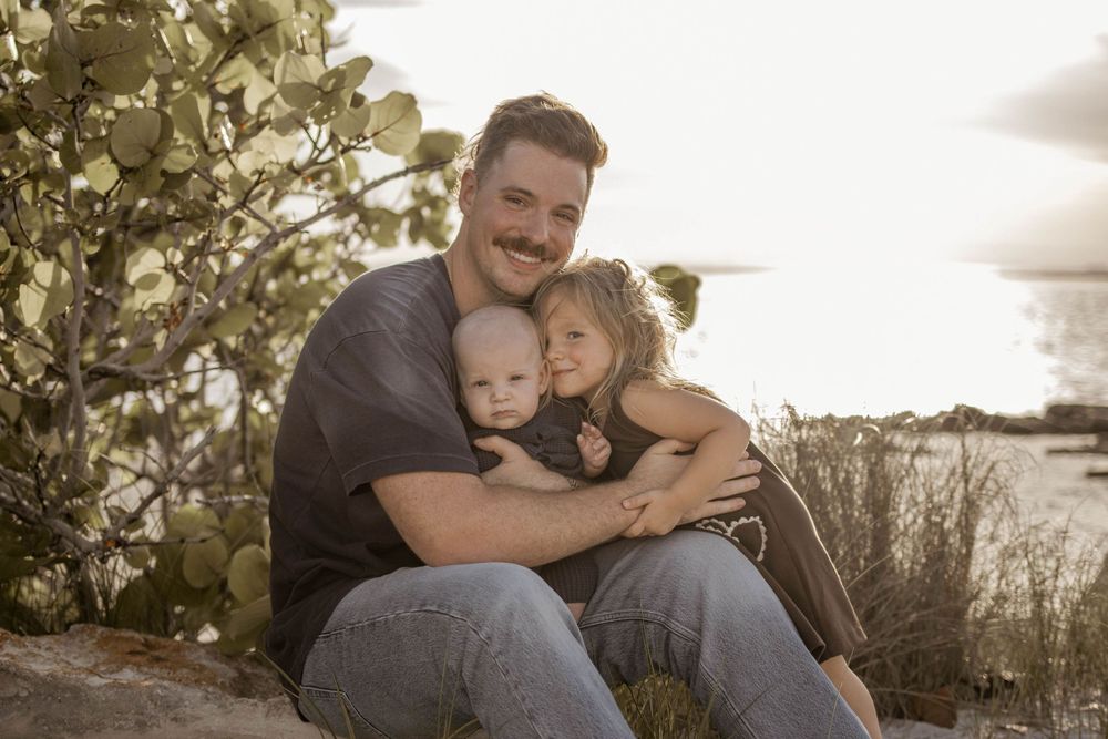 A man with a mustache sits outdoors hugging two young children near a cactus and water backdrop.