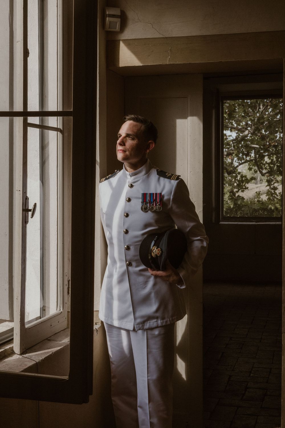 Military groom in white dress uniform with medals holds cap by sunlit window.