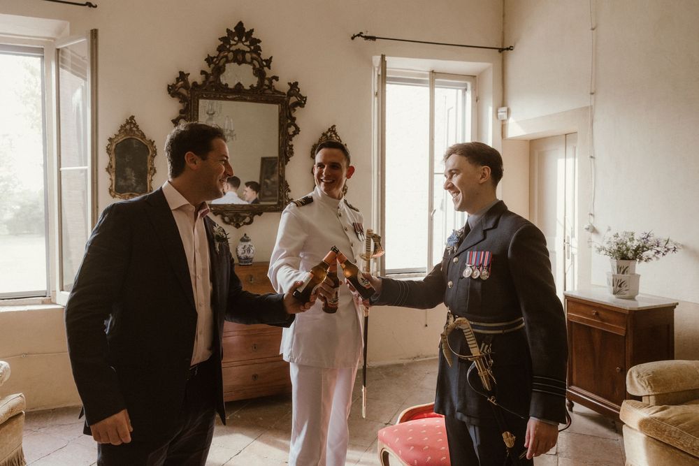 Military groom in dress uniform shares a joyful moment with two guests in an elegant vintage room before wedding ceremony.