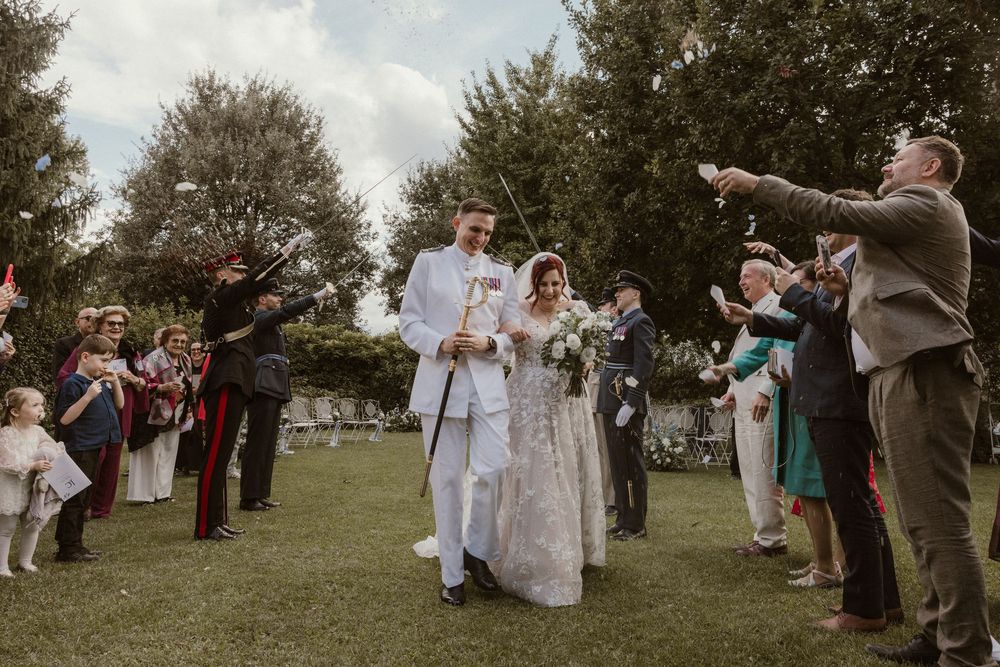 Bride and groom walk through confetti celebration on garden lawn surrounded by cheering wedding guests.