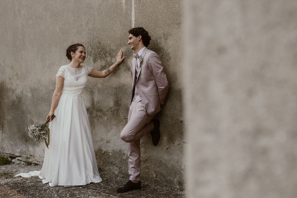 Bride in white dress and groom in pink suit pose against rustic concrete wall during outdoor wedding.