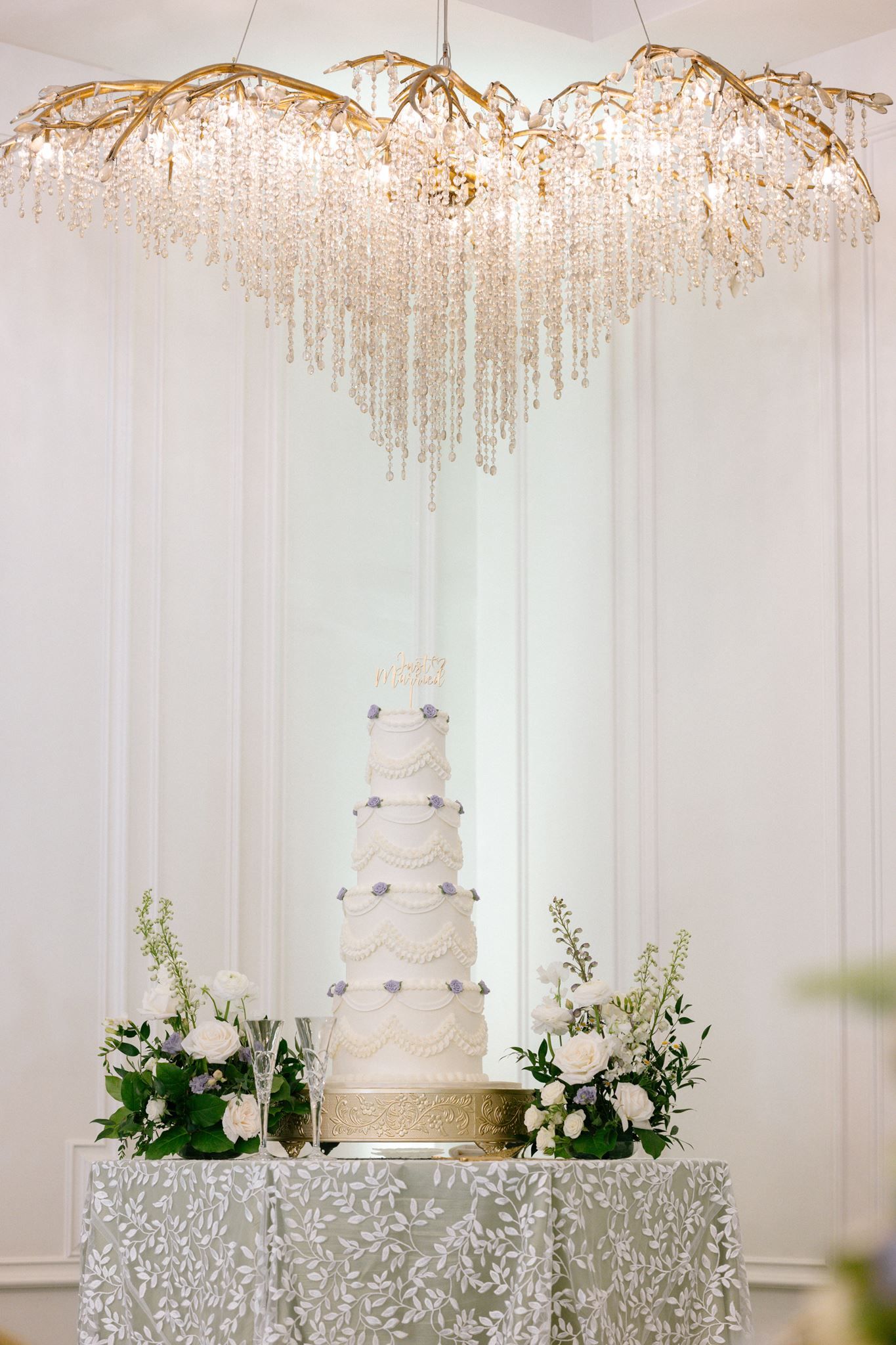 Elegant five-tier white wedding cake with floral accents displayed under a crystal chandelier on a silver embroidered table.
