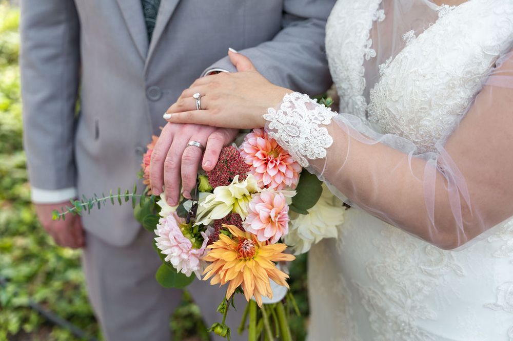 Bride and groom holding a colorful wedding bouquet with pink and orange flowers, showing wedding rings on their hands.