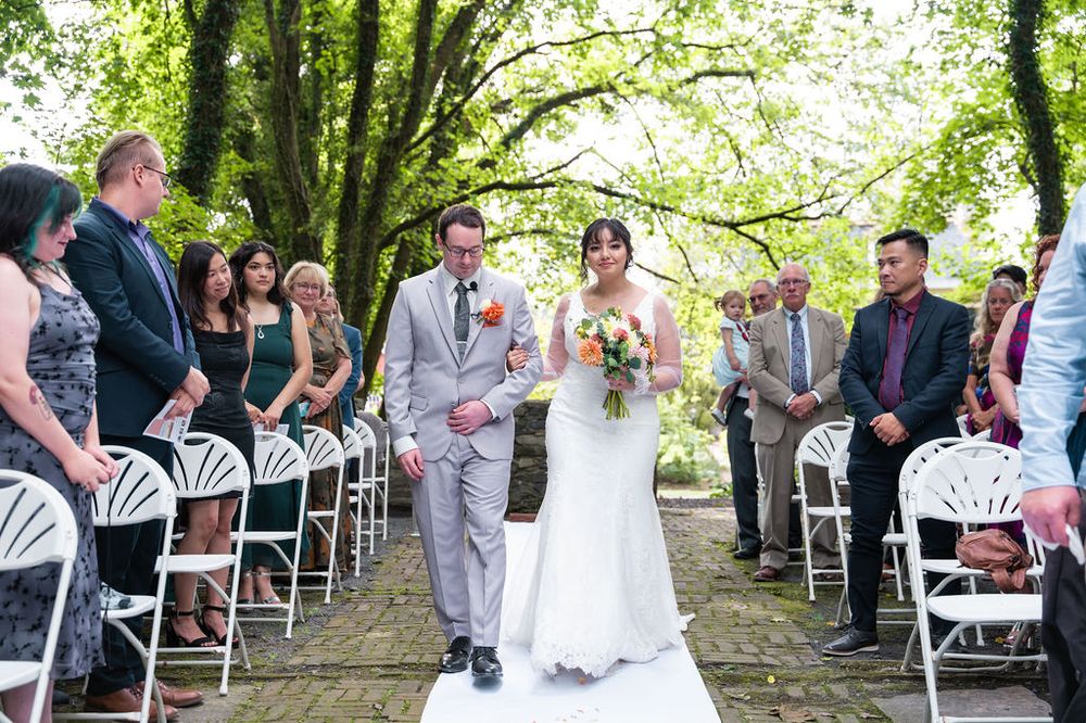 Bride and groom walk down outdoor wedding aisle lined with white chairs and guests under lush green trees.