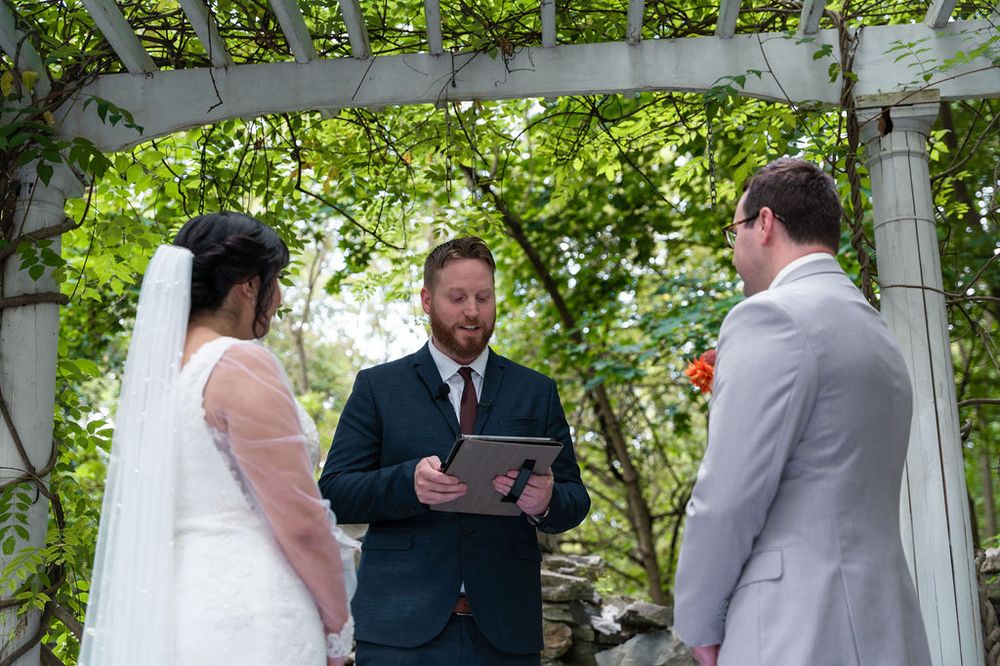 Outdoor wedding ceremony under a white arbor with lush greenery, officiant reading vows to bride in white gown and groom in gray suit.
