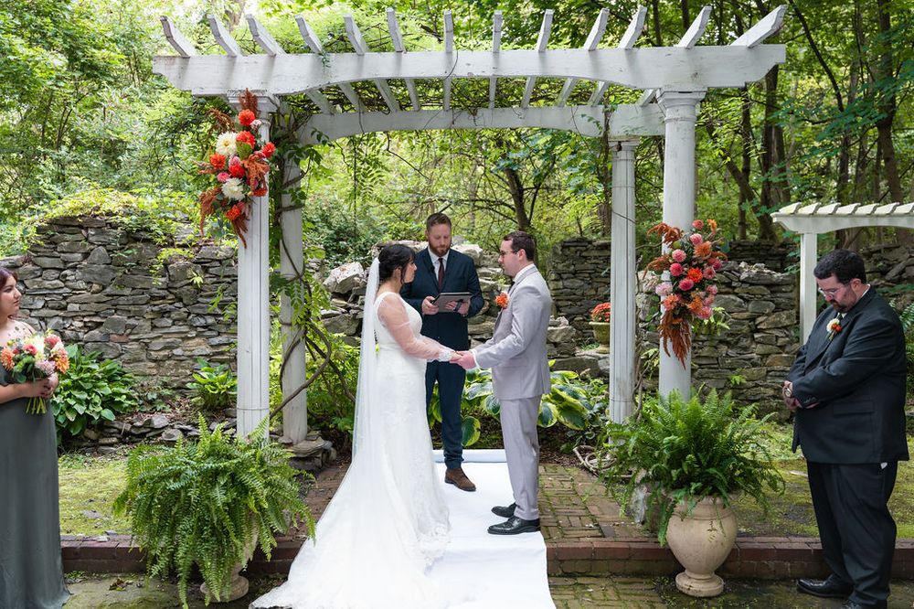Outdoor wedding ceremony under white pergola with floral arrangements, bride and groom exchanging vows with officiant.