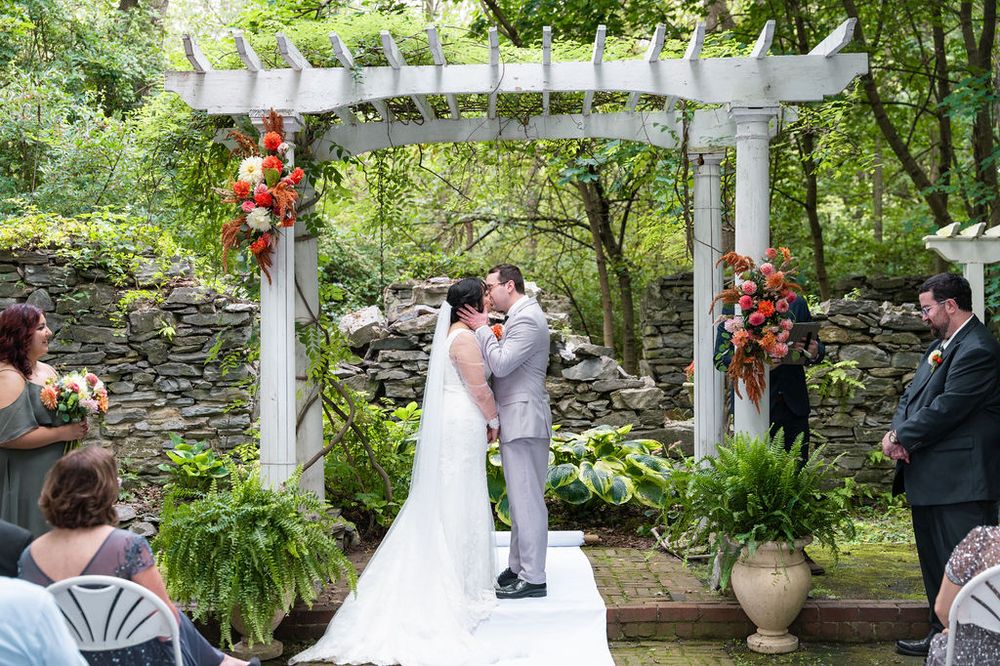 Couple shares first kiss under white pergola at outdoor garden wedding ceremony surrounded by lush greenery and floral arrangements.