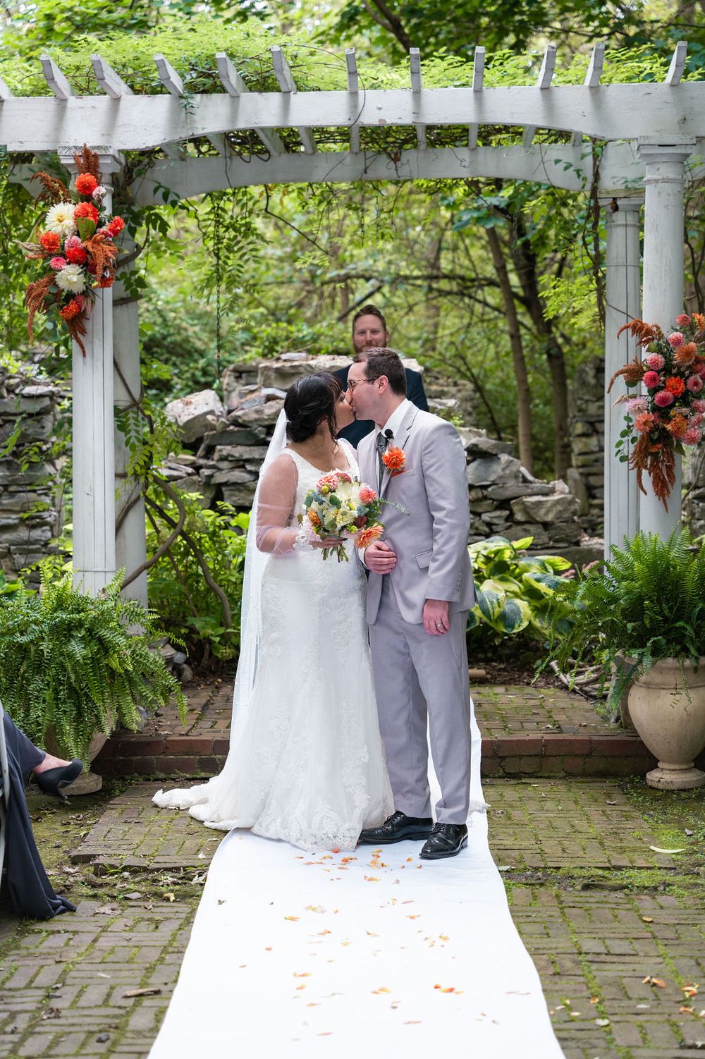 Bride and groom share first kiss under white pergola with orange floral arrangements at outdoor garden wedding ceremony.