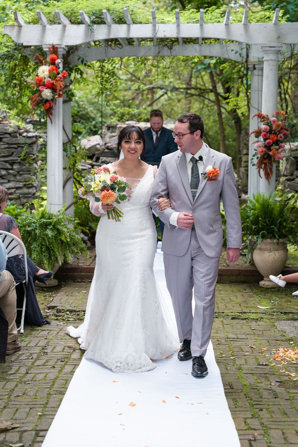 Bride and groom walk down white aisle after outdoor garden wedding ceremony under white pergola.