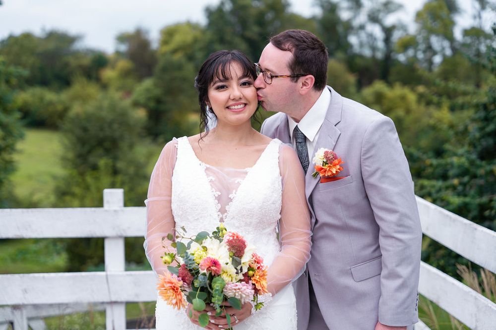 Bride holding colorful bouquet smiles as groom in gray suit kisses her cheek near white fence outdoors.