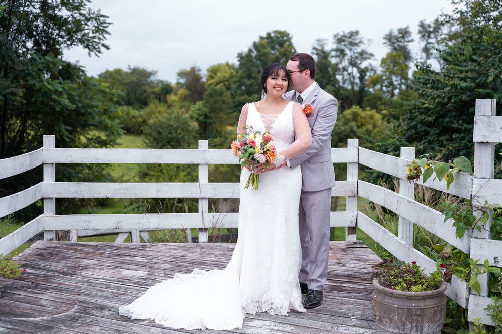 Bride and groom in gray suit pose together on white wooden deck surrounded by lush green trees.