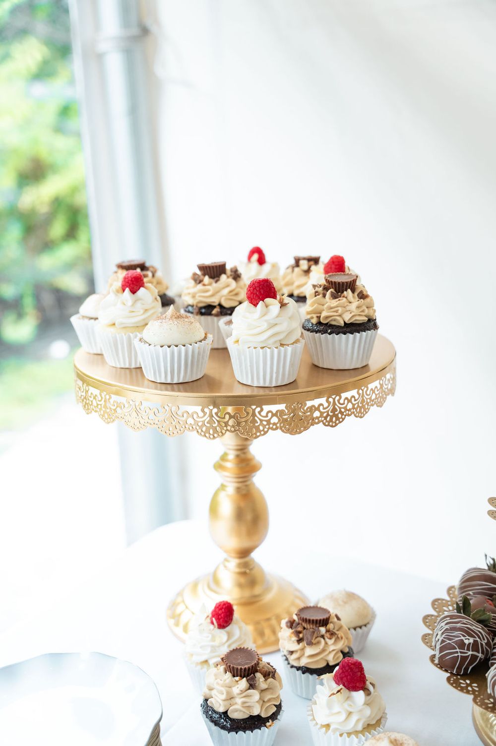 Gold pedestal stand displaying elegant cupcakes topped with cream frosting, raspberries, and chocolate candies at a wedding dessert table.