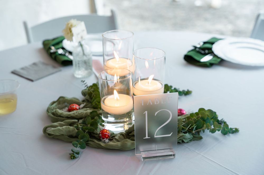 Elegant wedding table centerpiece with floating candles in glass vases, greenery, and a frosted acrylic table number 12.