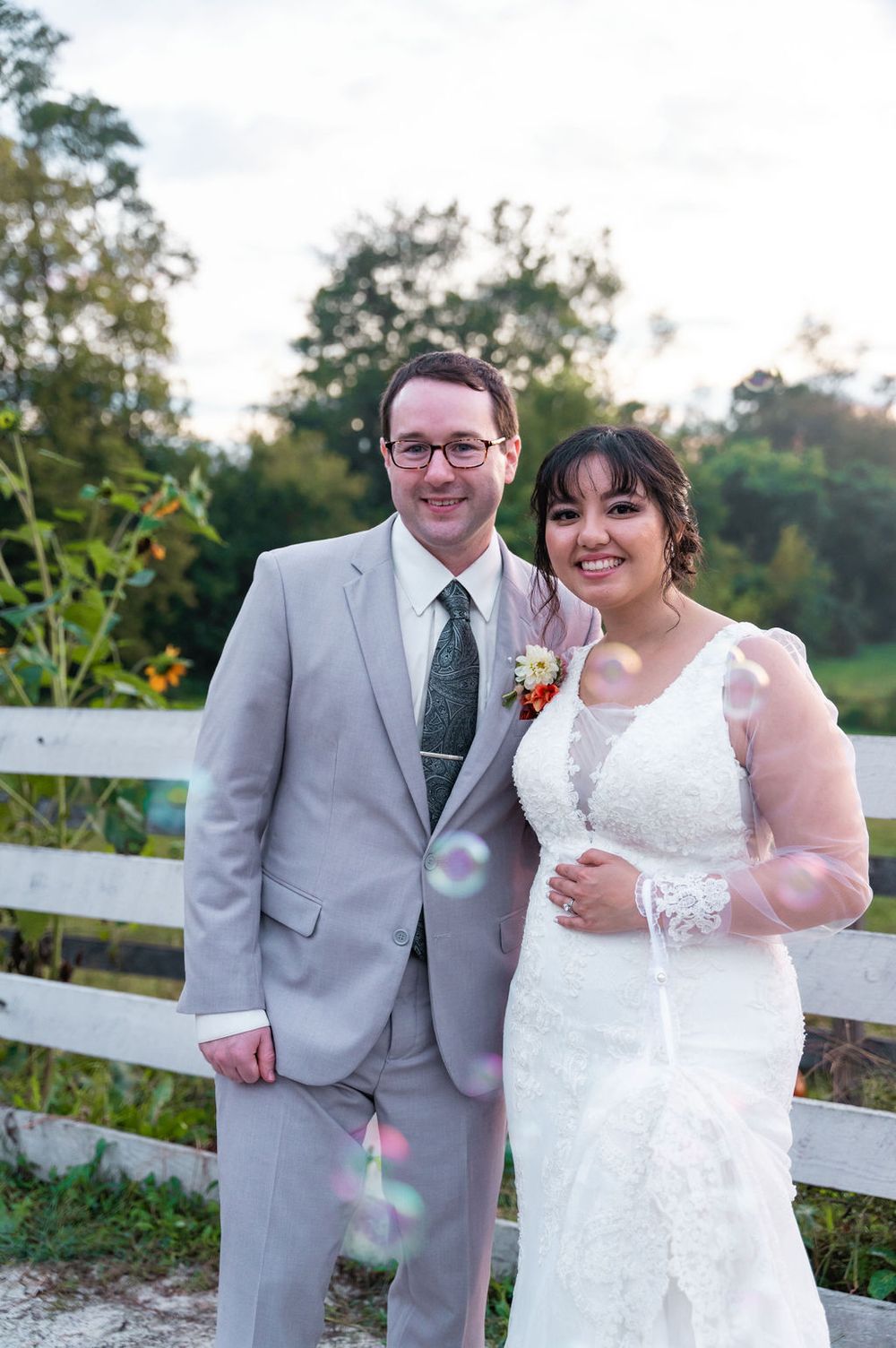 Bride in lace gown and groom in gray suit pose together by white fence at outdoor wedding ceremony.