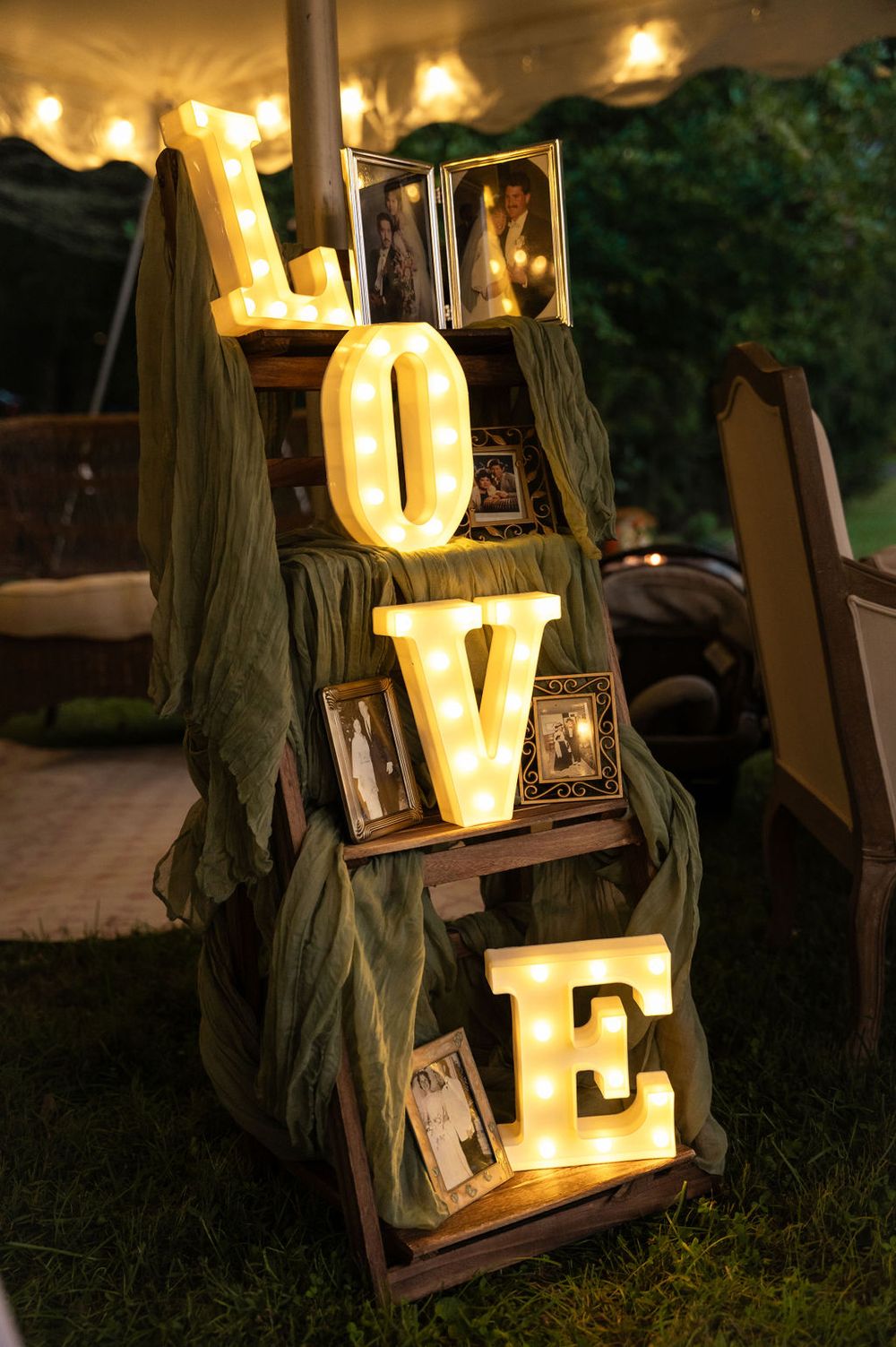 Illuminated marquee LOVE letters on rustic wooden ladder with framed photos and green fabric at outdoor wedding reception.